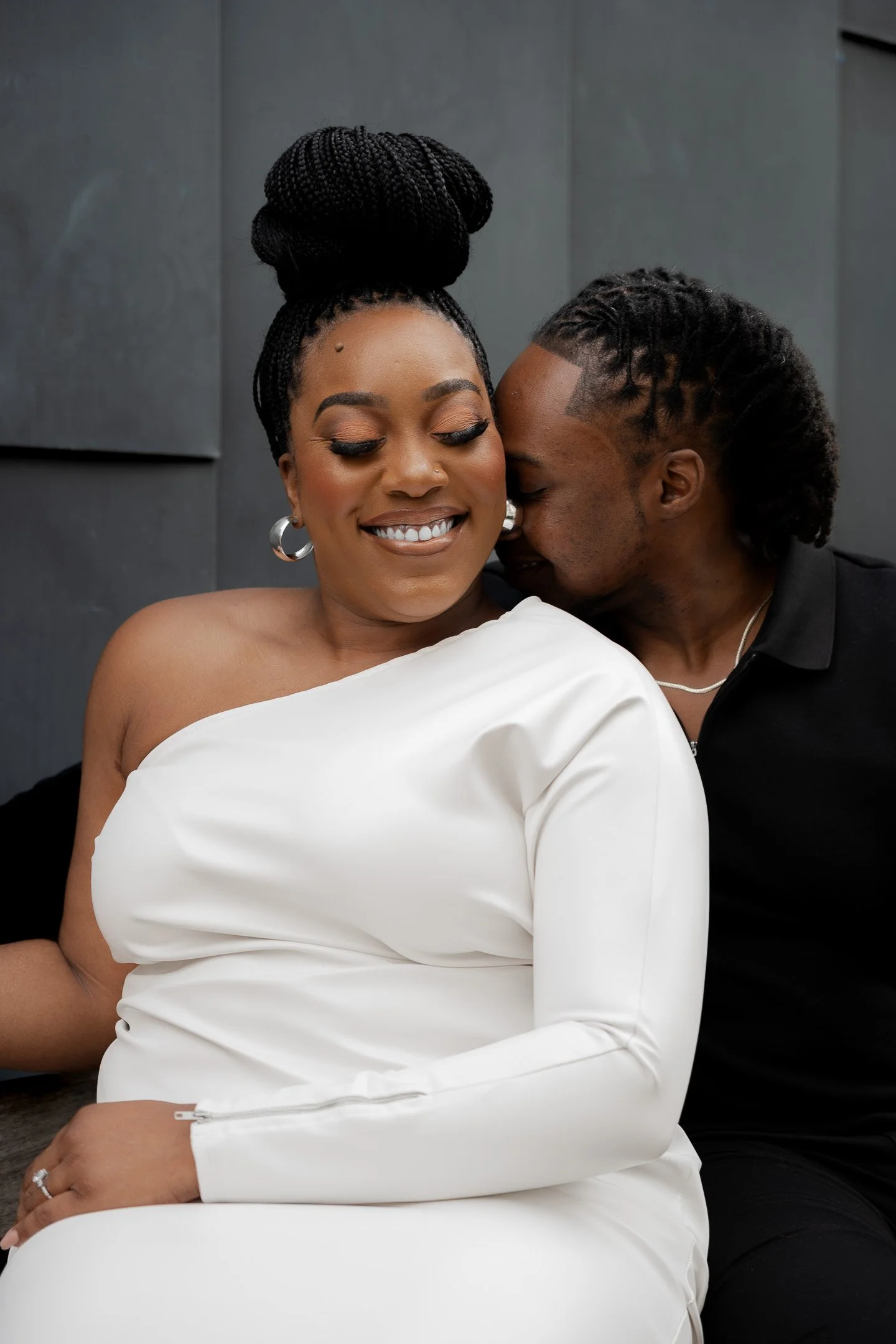 A smiling woman in a white dress sitting next to a man in a black outfit, who is leaning in closely, appearing affectionate.