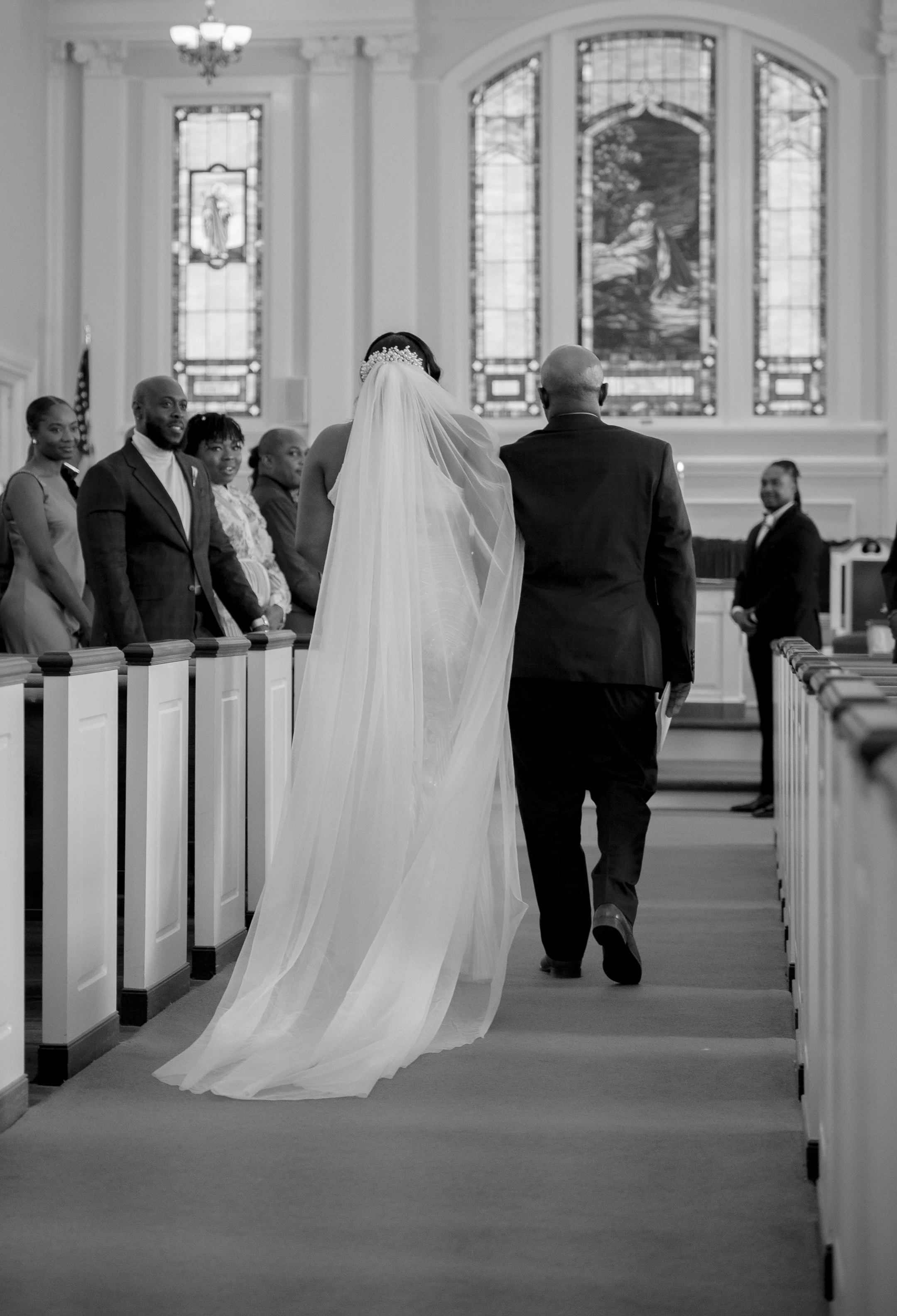 A bride and a man, possibly her father, walking down the aisle of a church during a wedding ceremony. The bride is wearing a long veil and there are guests seated on either side. The groom or officiant waits at the altar. The church features stained 