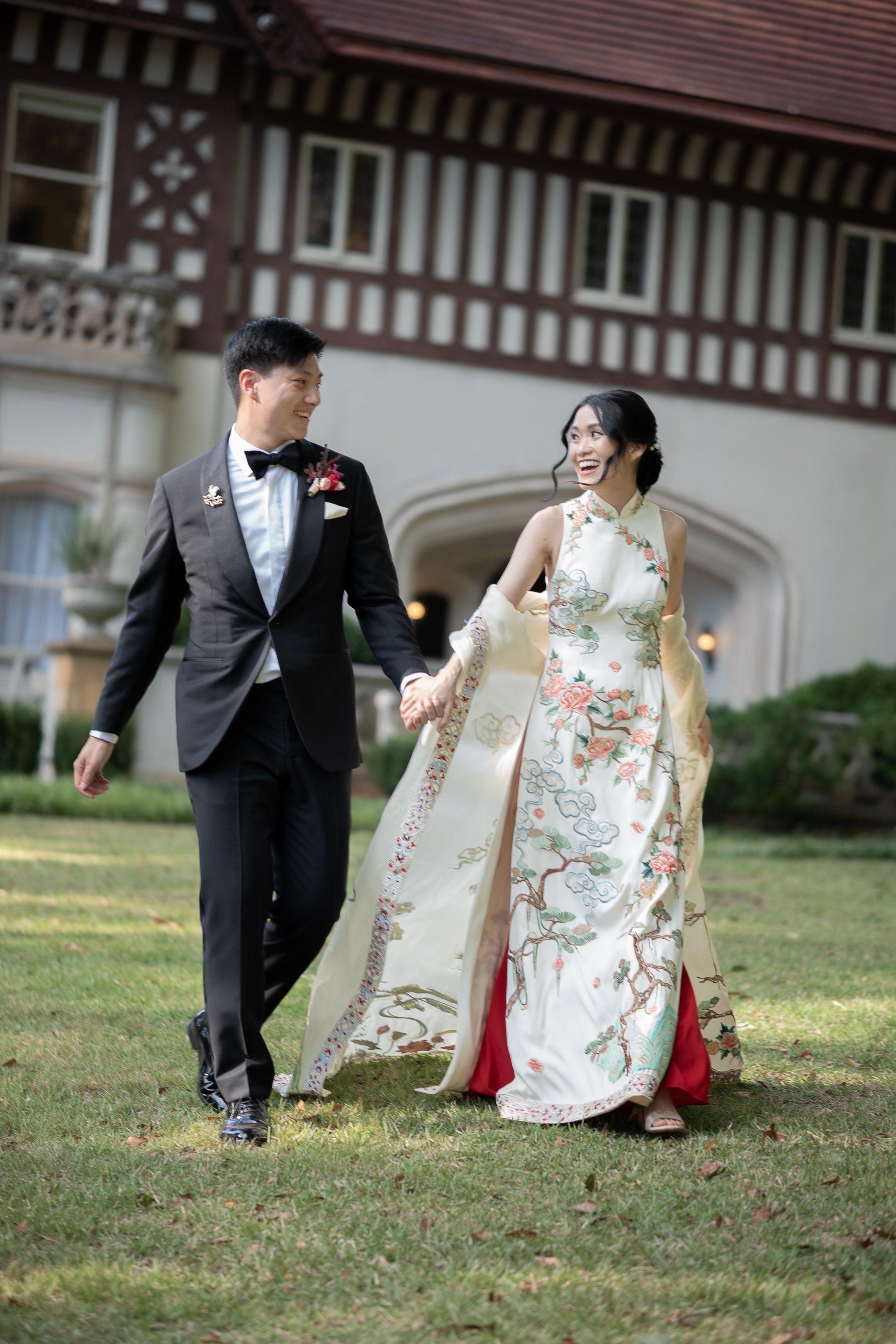 A newlywed couple, with the groom in a black tuxedo and the bride in a traditional Asian wedding dress, holding hands and smiling as they walk on a lawn outside a historic building.