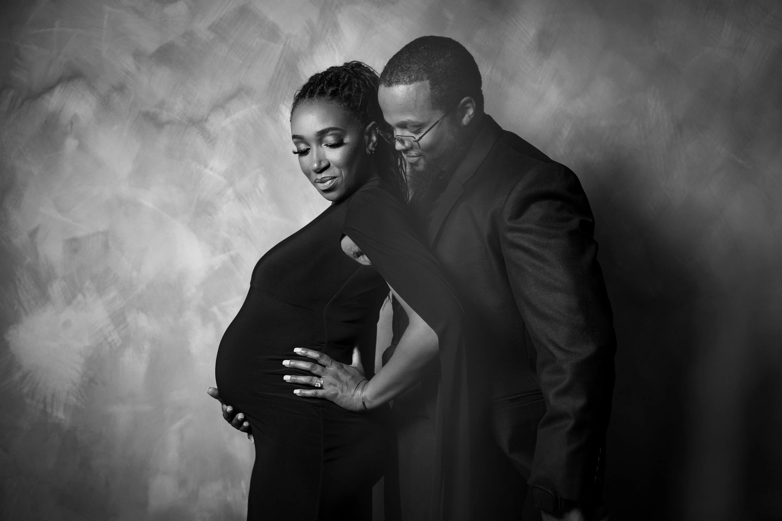 A pregnant woman in a black dress with a man in a suit standing behind her, both looking down and touching her belly, in a black-and-white photo.