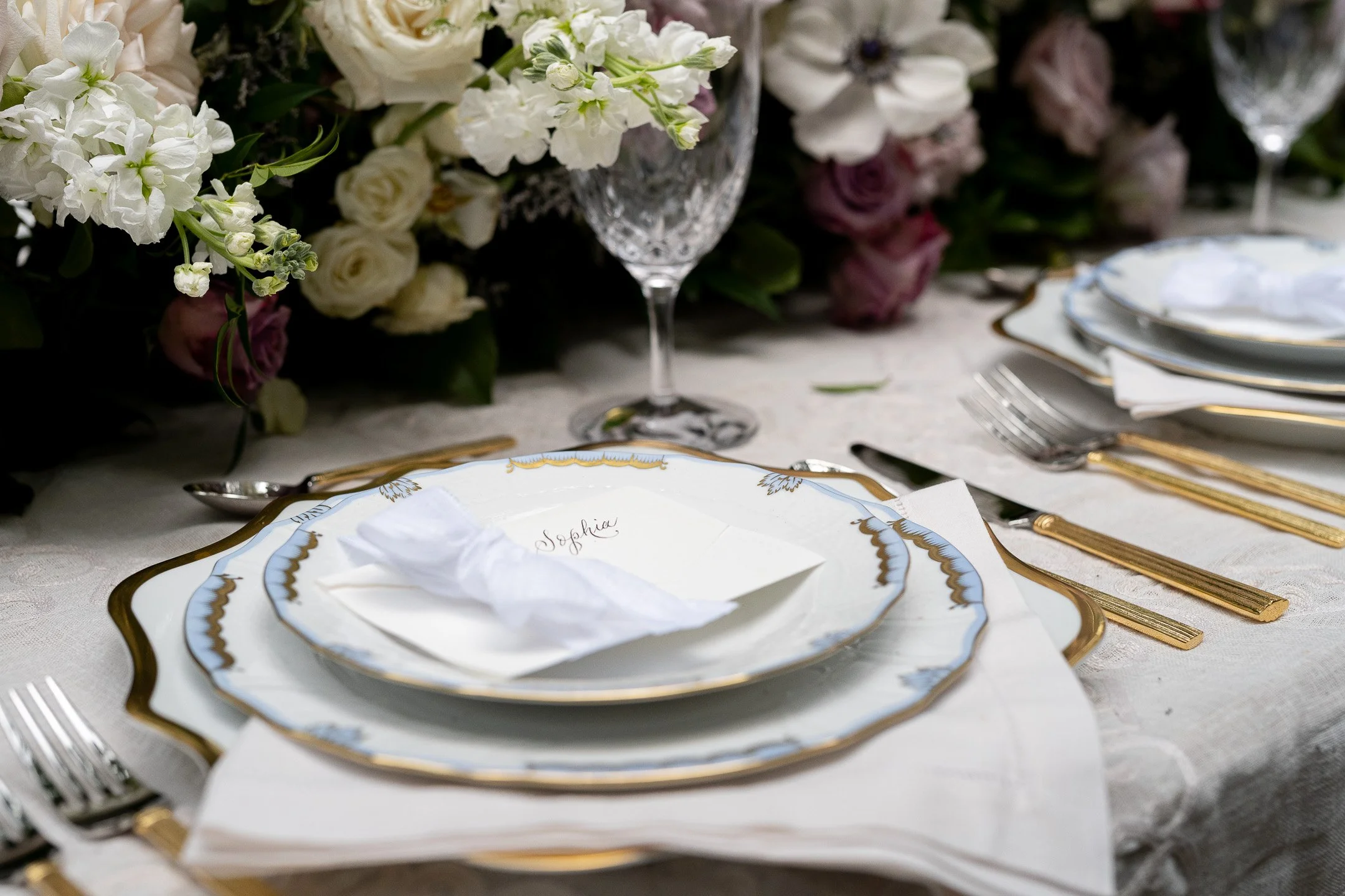 Elegant table setting with decorative plates, gold cutlery, and a crystal glass, accompanied by floral centerpiece.