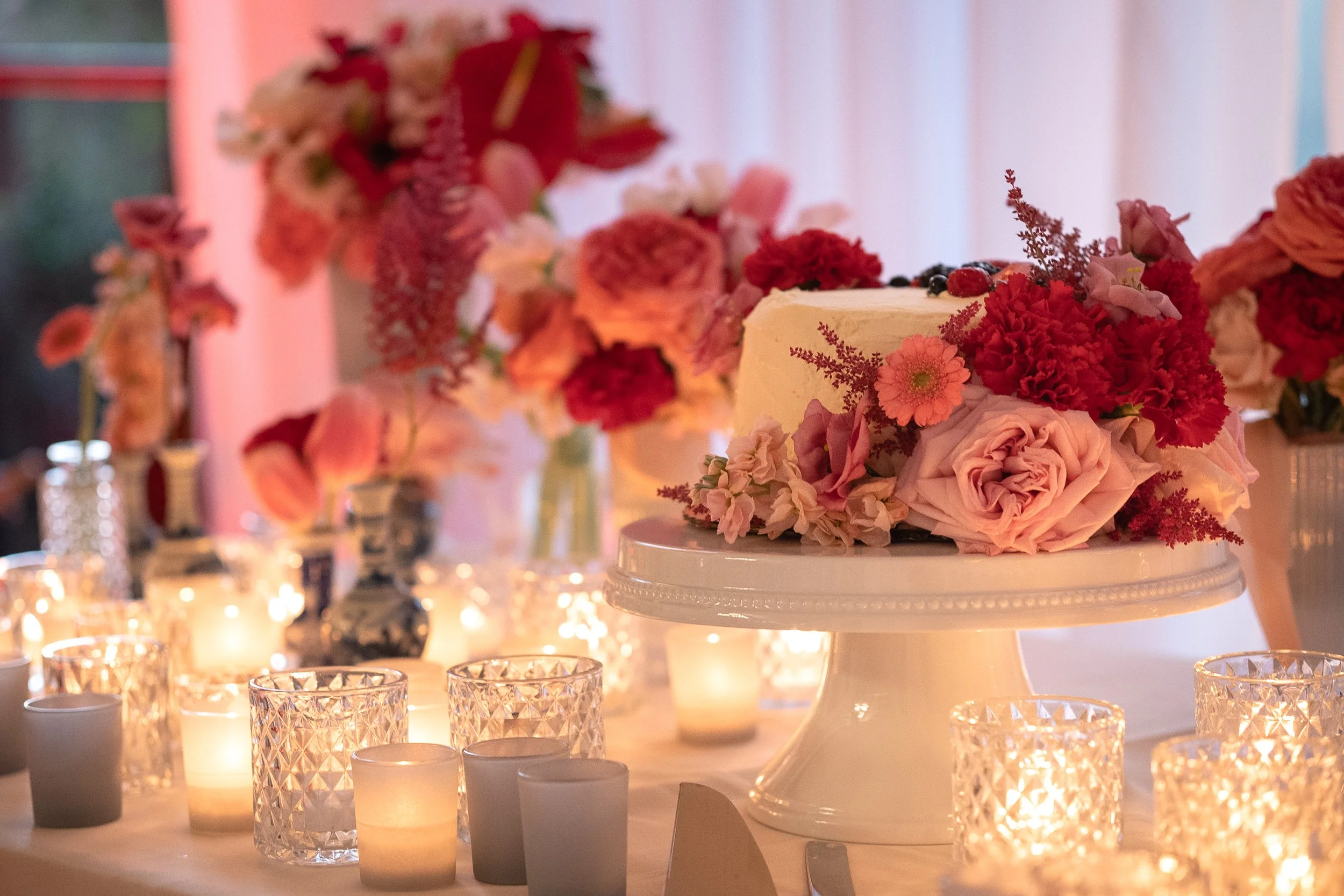 A wedding cake decorated with pink and red flowers surrounded by candle votives on a table with pink floral arrangements and soft lighting.
