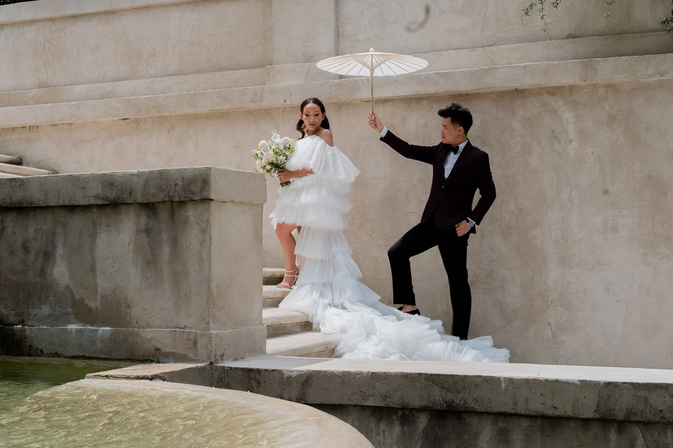 A wedding couple on stone steps, the bride in a tiered white gown holding a bouquet, the groom in a dark suit holding a white parasol.
