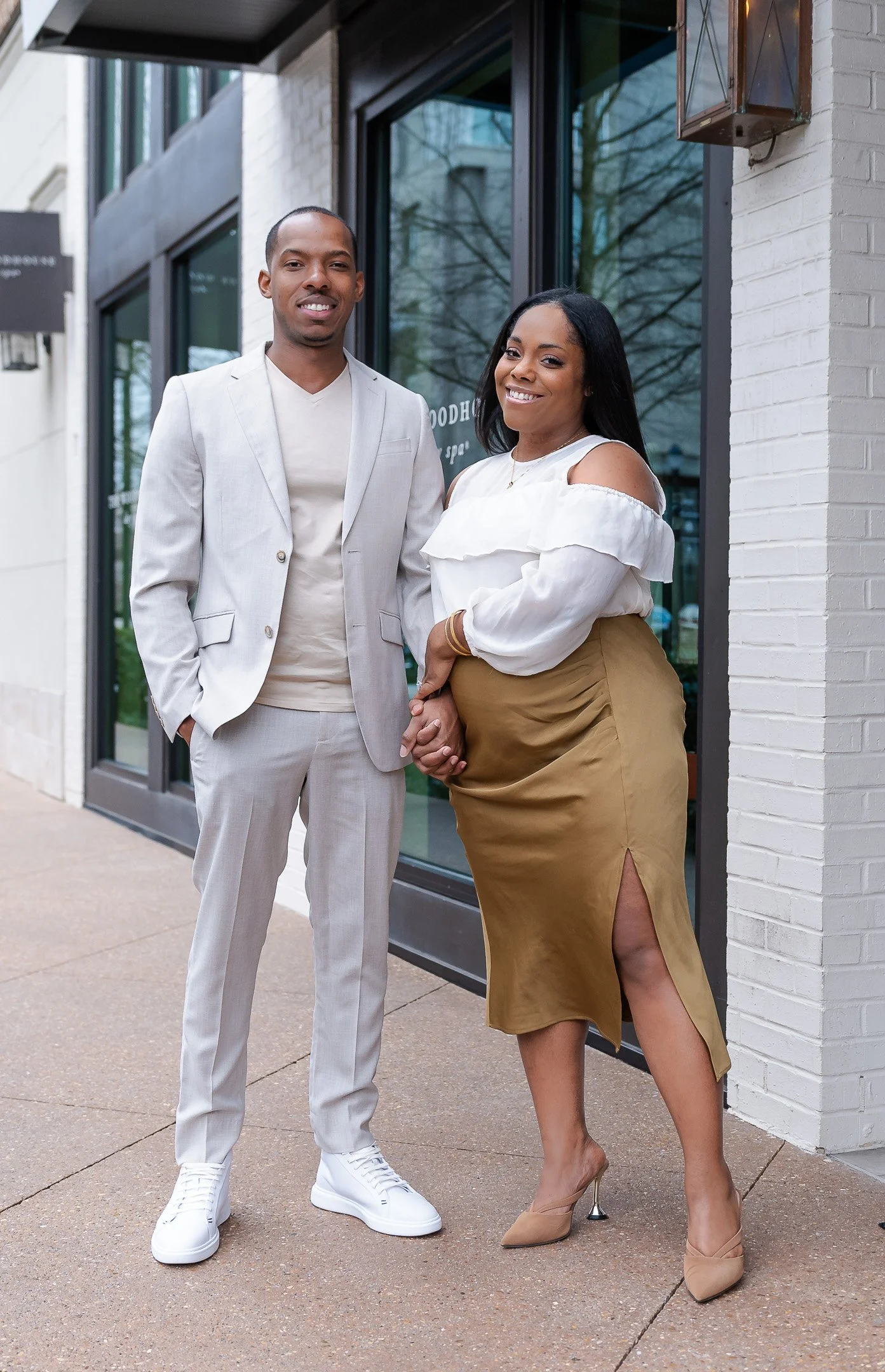 A smiling couple stands hand in hand outside a modern building. The man wears a light gray suit and white sneakers, while the woman wears a white ruffled blouse and a tan skirt.