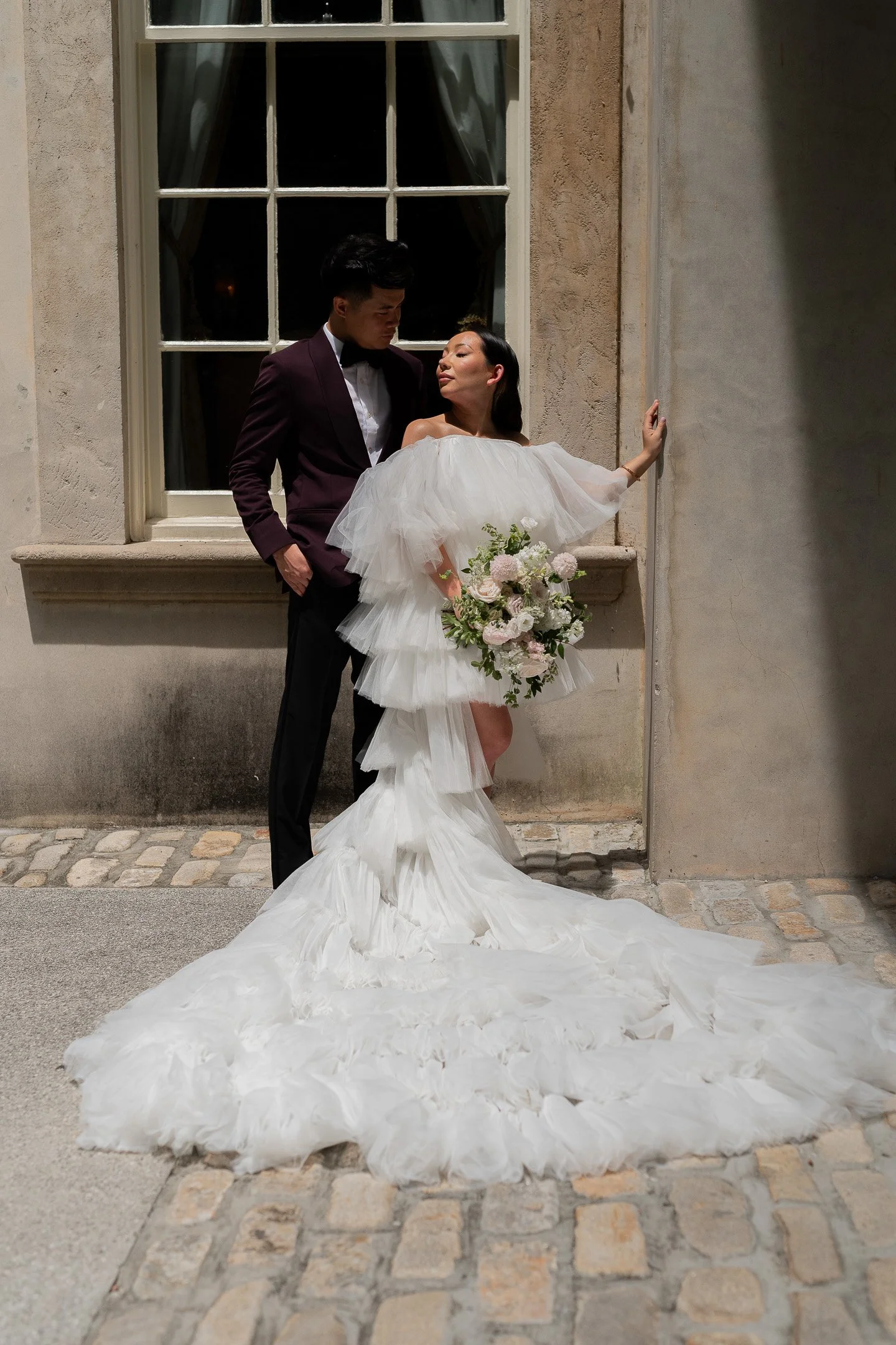 Bride in ruffled white dress holding floral bouquet stands beside groom in dark suit, posing against an outdoor wall in sunlight.