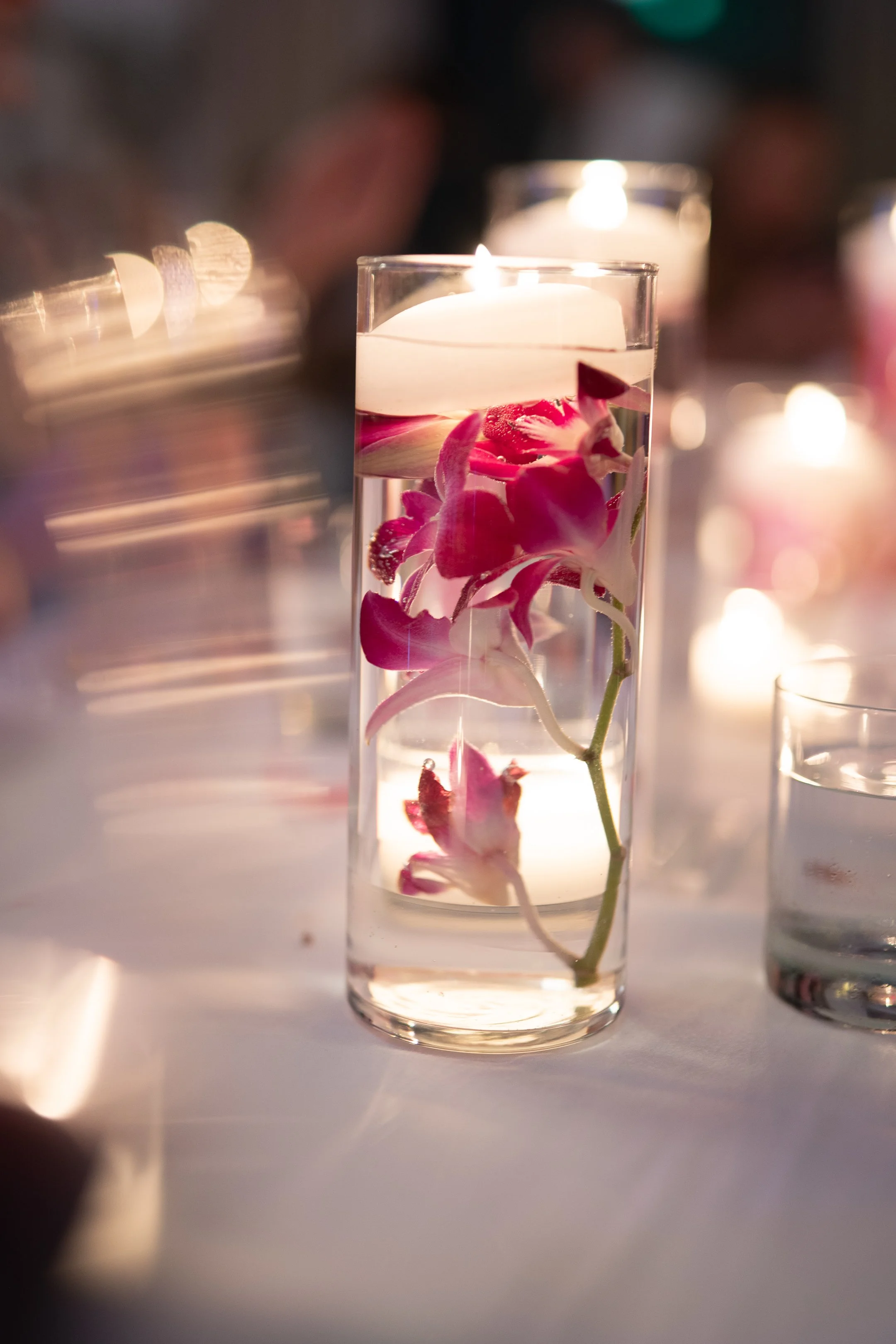 A tall glass vase with a floating candle and pink orchids submerged in water, placed on a white table with blurred candlelit background.