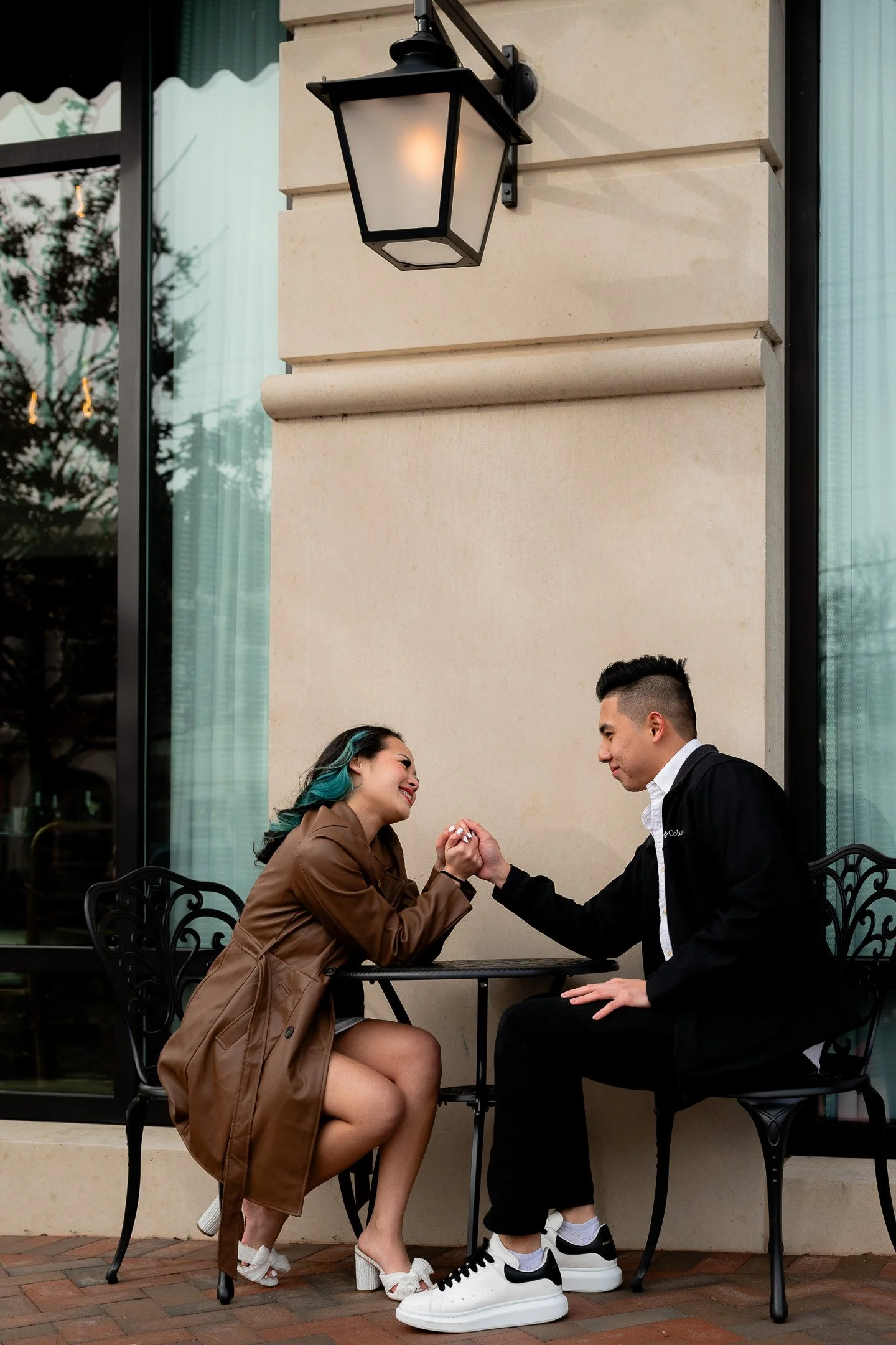 A couple sitting at an outdoor cafe table, holding hands and smiling. The woman is wearing a brown coat and white shoes, while the man is in a black jacket and white sneakers. They are seated under an exterior wall light with a window in the backgrou