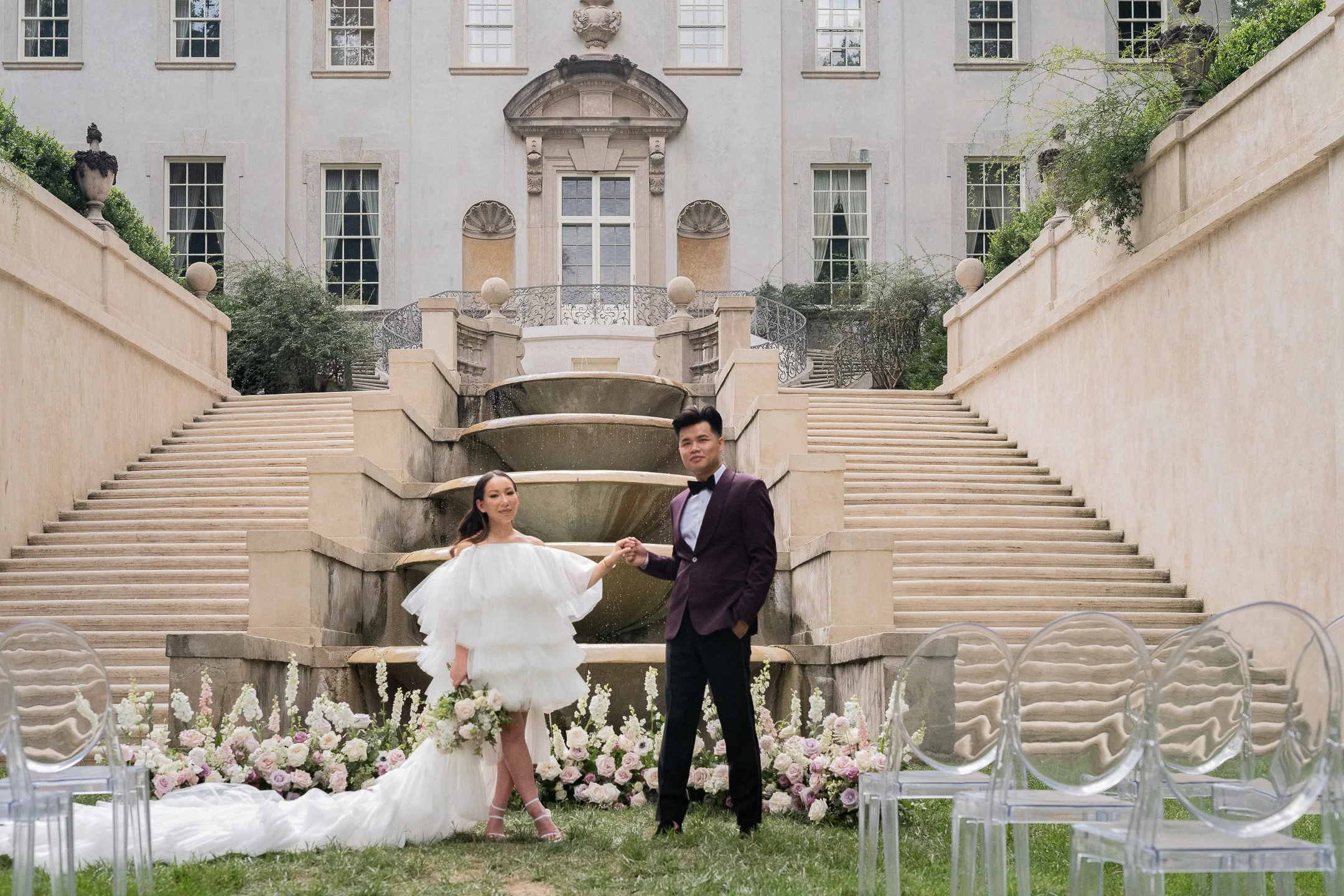 Bride and groom standing hand in hand in front of a historic mansion, with a fountain, staircase, and floral decorations.