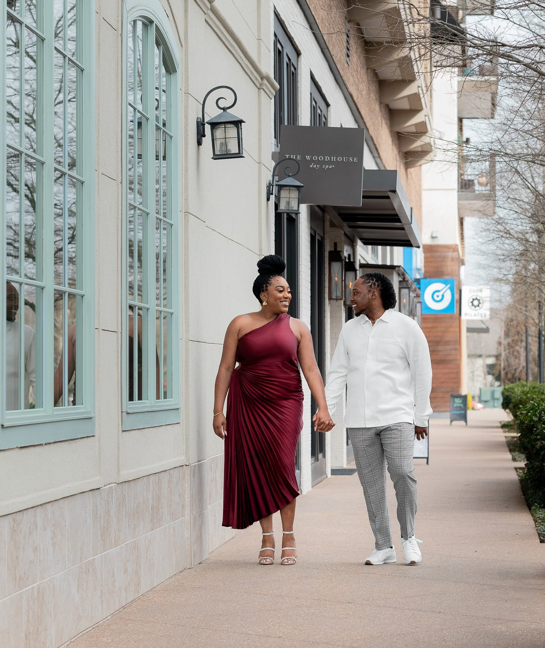 Couple walking happily holding hands on a sidewalk near a building with large windows and a sign for The Woodhouse Day Spa.