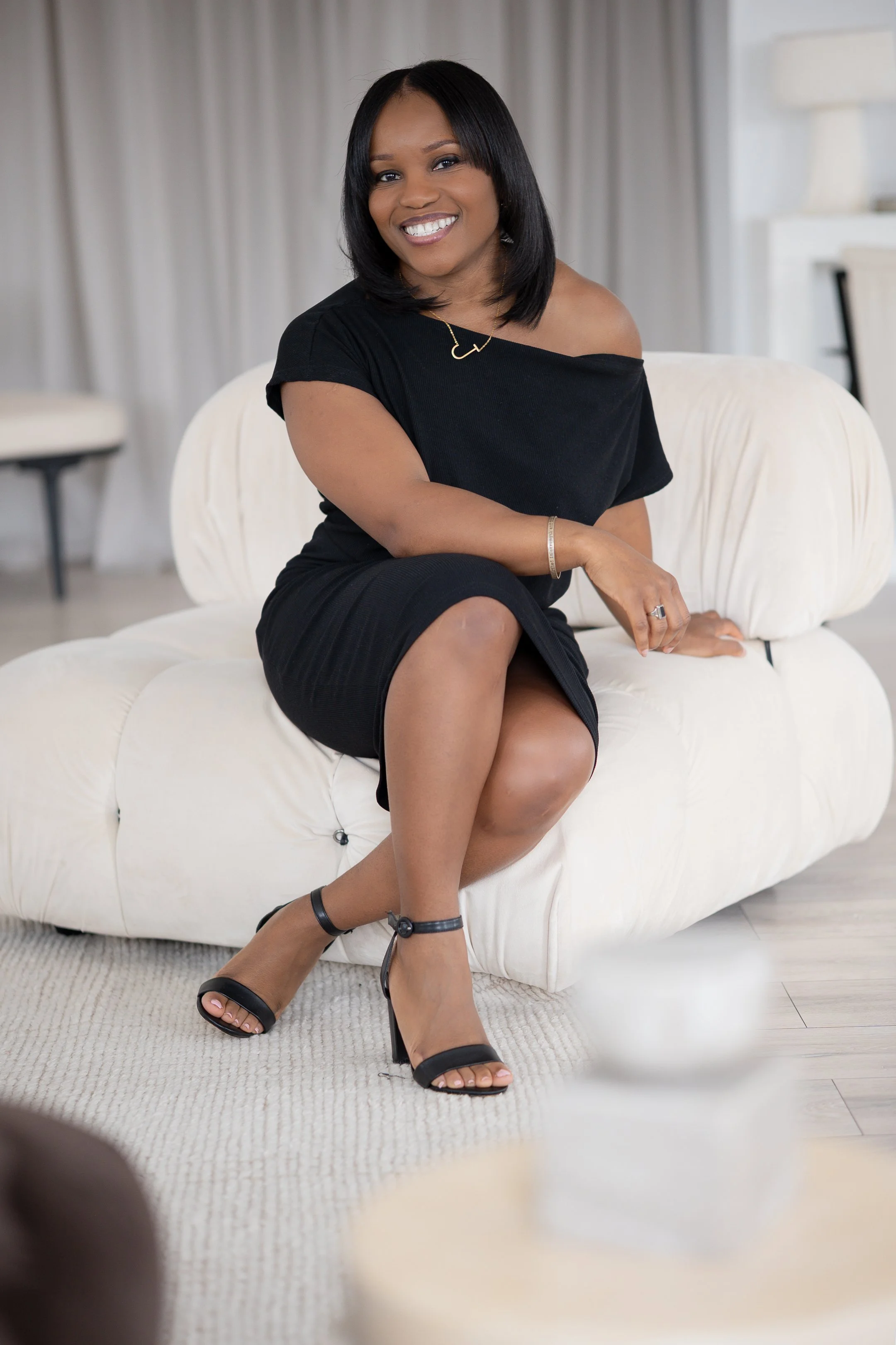 A woman in a black dress sitting on a white sofa, smiling at the camera. She is wearing black heeled sandals and a gold bracelet, with straight black hair. The room has a soft, neutral decor.
