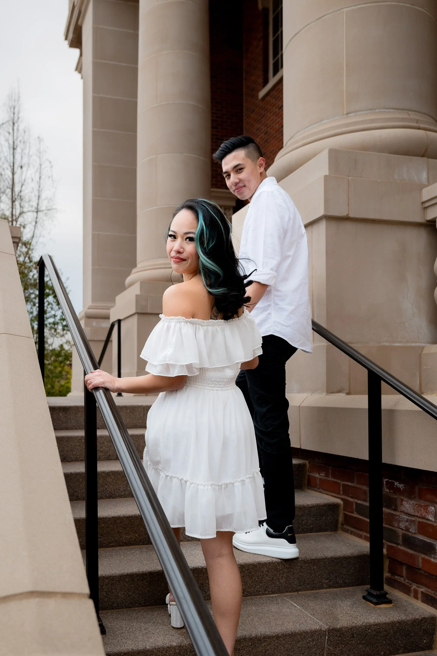 A woman in a short white off-the-shoulder dress and a man in a white shirt and dark pants are walking up a stone staircase outside a building with columns.