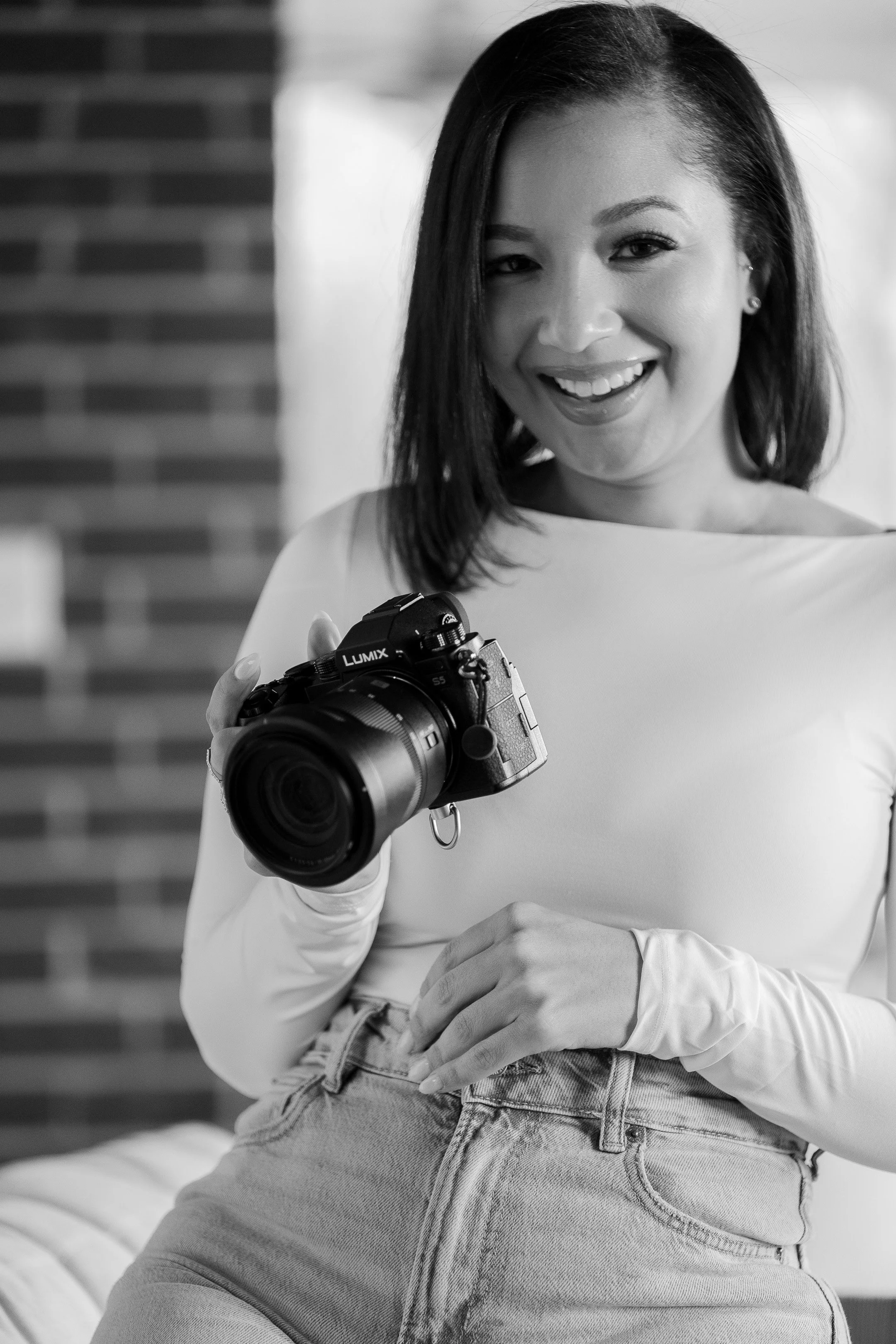 Smiling woman holding a Lumix camera, wearing a long-sleeve top and jeans, with a brick wall in the background, in black and white.