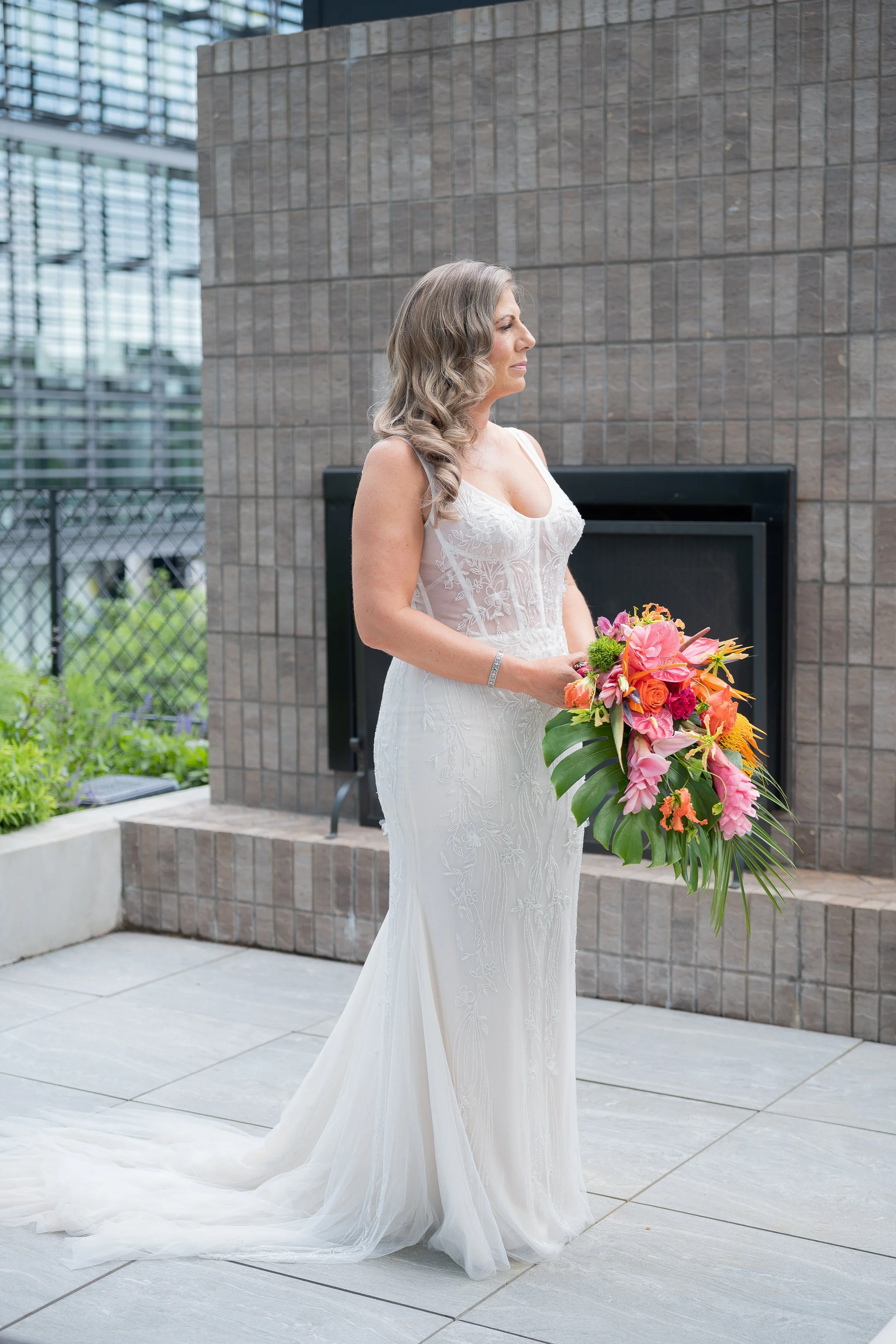A woman in a white wedding dress holding a colorful bouquet of flowers, standing outdoors near a brick wall and a fireplace.