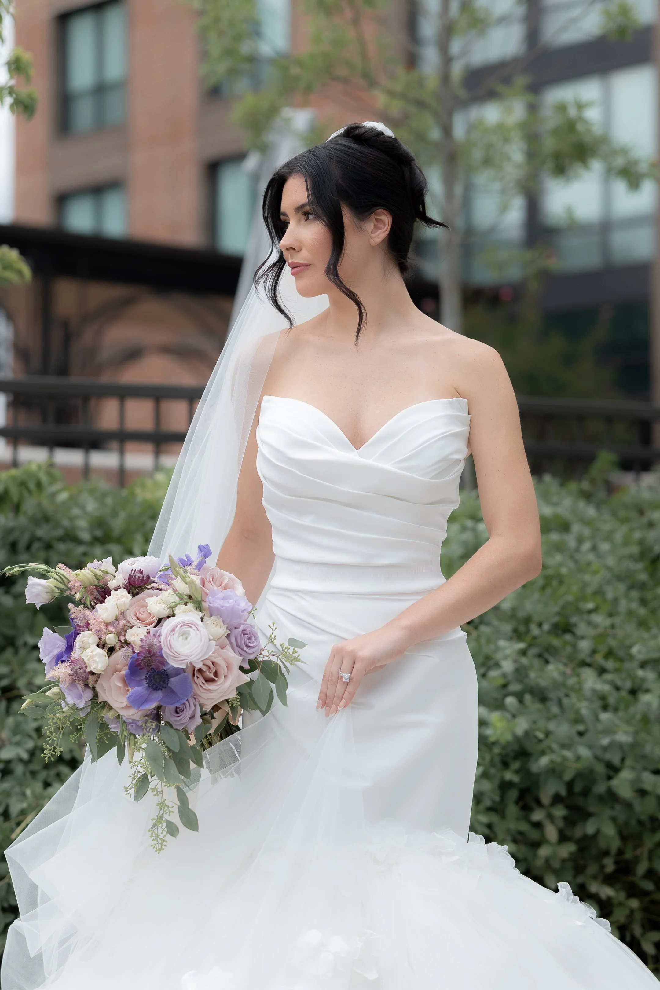 A bride in a strapless white wedding gown holding a bouquet of purple and pink flowers, standing outdoors with a modern building and greenery in the background.
