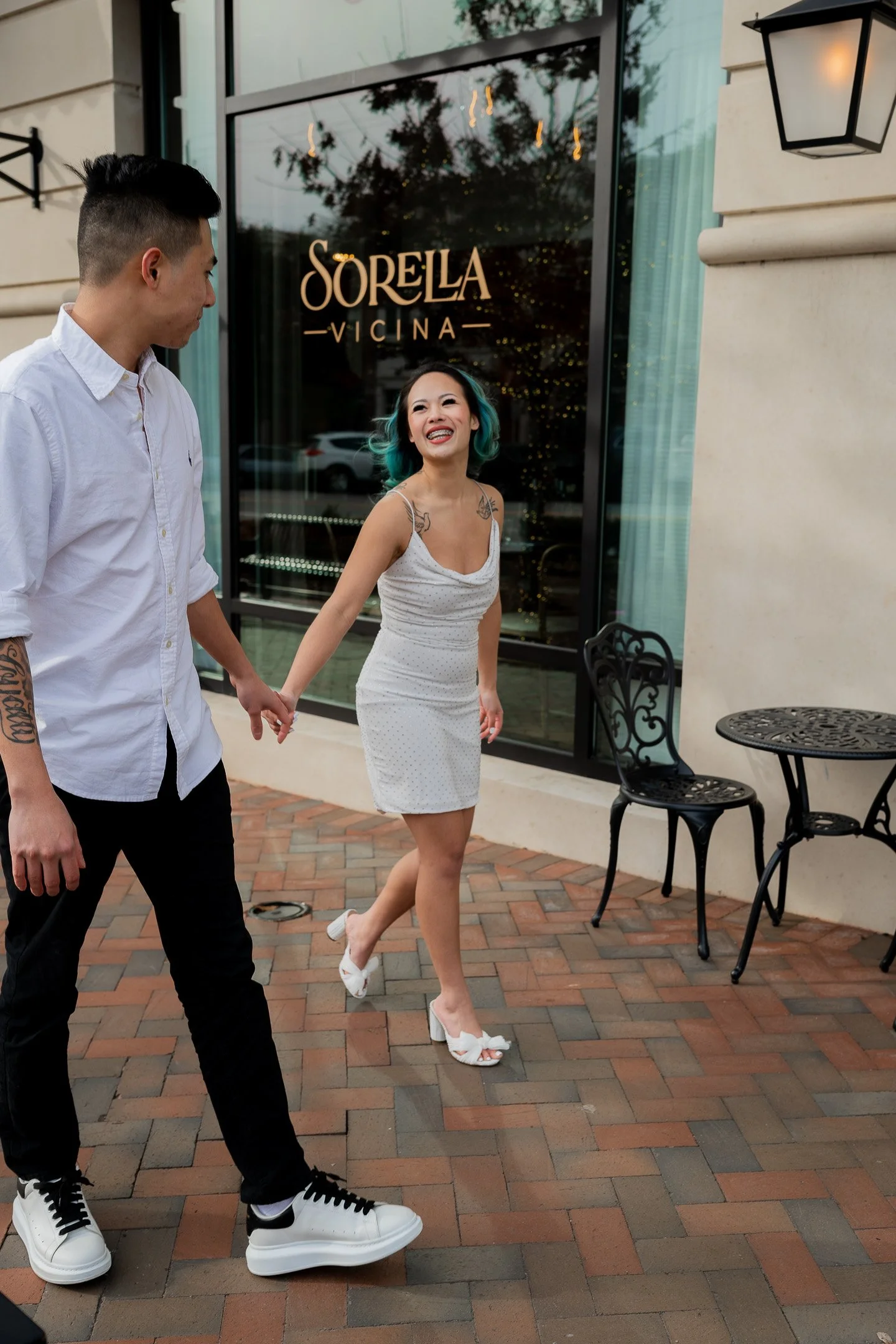 A couple walking outside a building labeled 'Sorella Vicina.' The woman is smiling and wearing a white dress, while the man wears a white shirt and black pants. They appear happy and are holding hands.