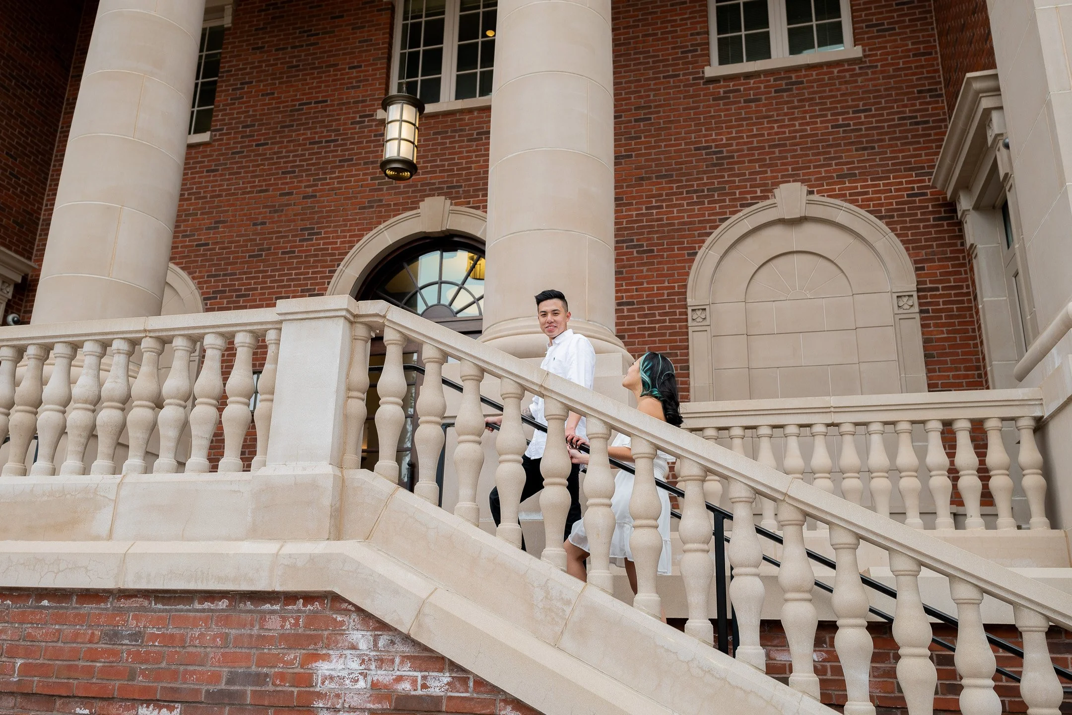 Two people walking up an outdoor staircase with ornate stone railings and brick building in the background.