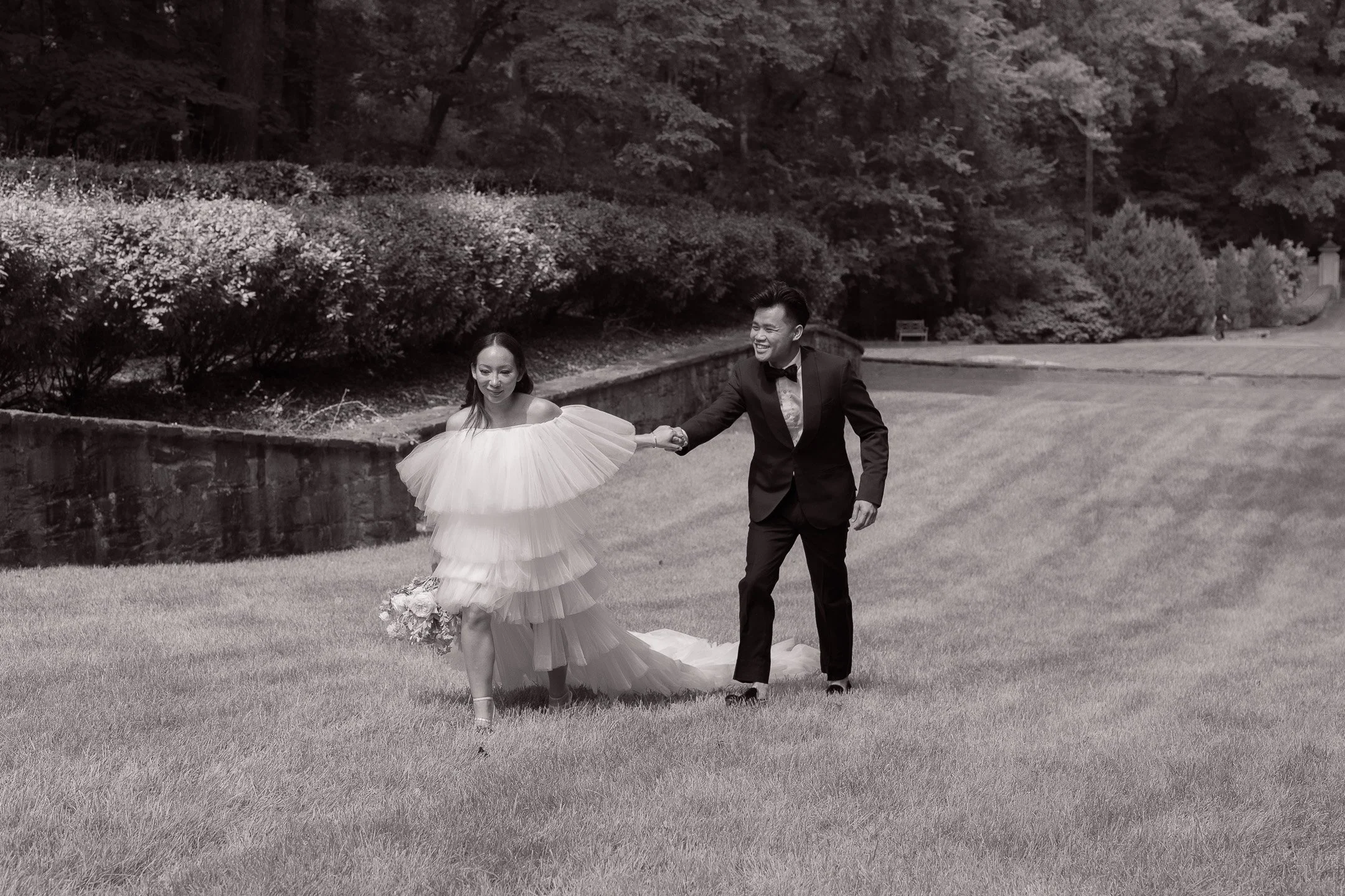 A couple in formal attire walking on a grass lawn, with the woman in a tiered tulle dress holding a bouquet, and the man in a suit and bow tie, both smiling and holding hands, in a garden setting.