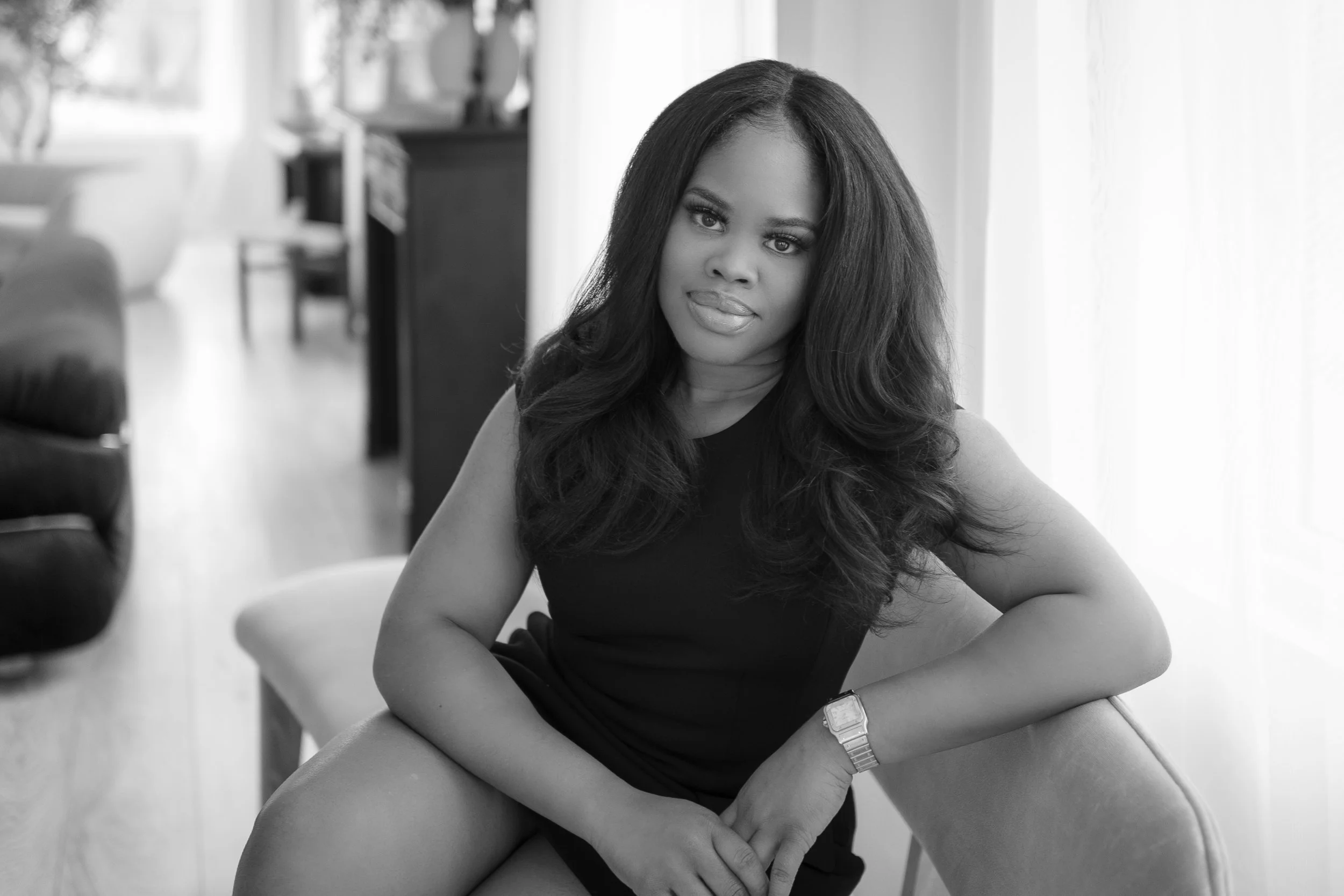 Black and white portrait of a woman in a black dress seated on a chair, with long hair, against a softly lit indoor background.