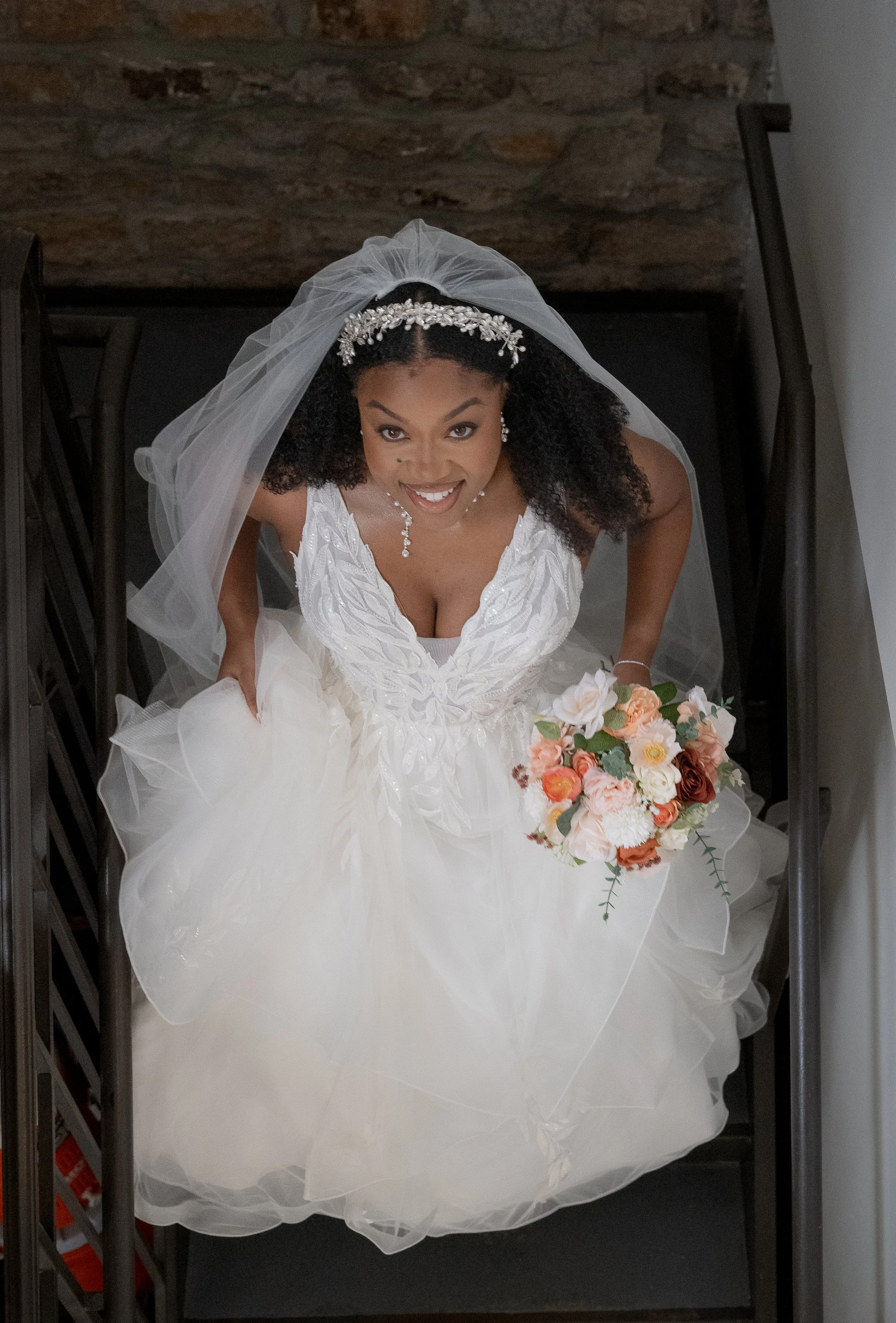 Bride wearing a white wedding dress and veil, holding a bouquet of flowers, and looking up while standing on a staircase.
