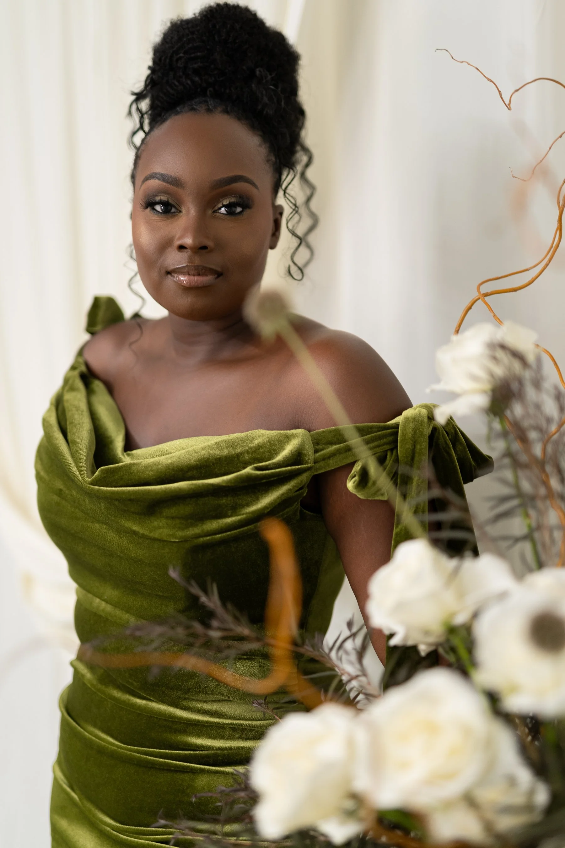 Woman wearing a green velvet dress, standing next to white flowers and branches. The background is light-colored.