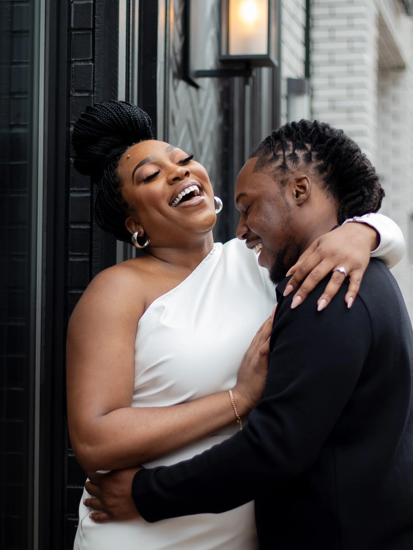A couple embracing and smiling, with the woman wearing a white dress and the man in a dark shirt, standing outdoors near a black and white building.