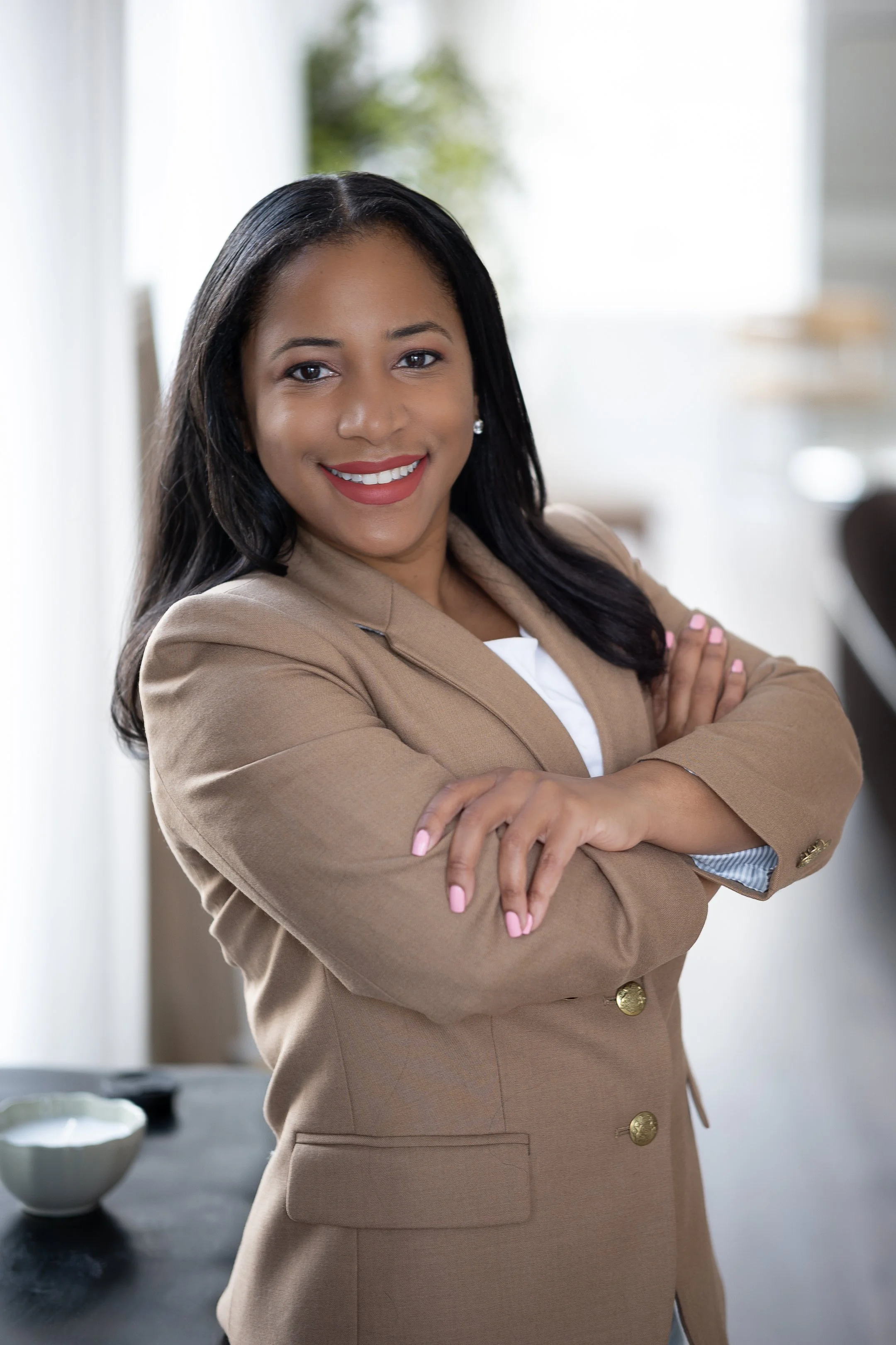 Smiling person with long hair wearing a beige blazer, arms crossed.