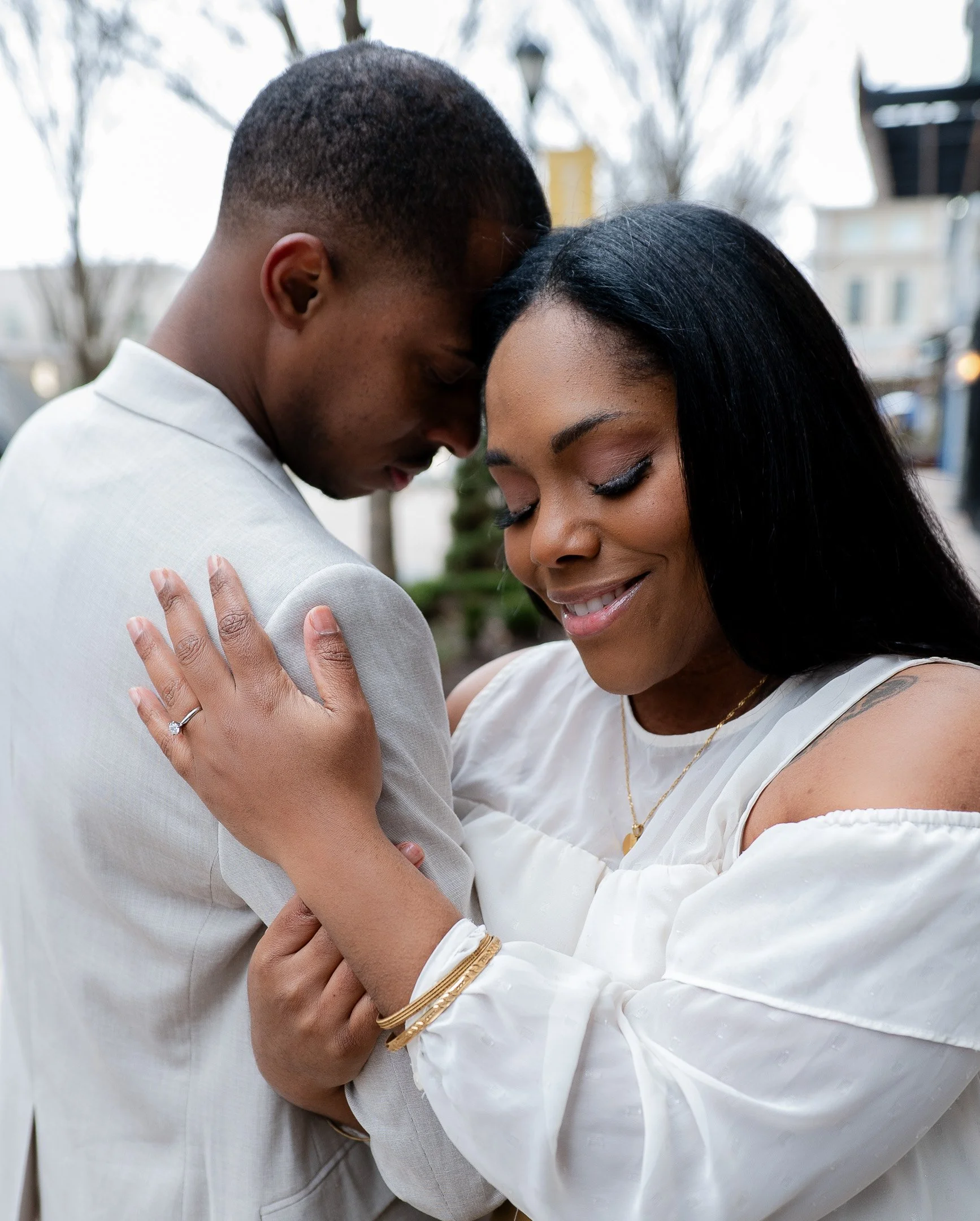 A couple embracing, with a woman showing an engagement ring on her hand, outdoors in a city setting.