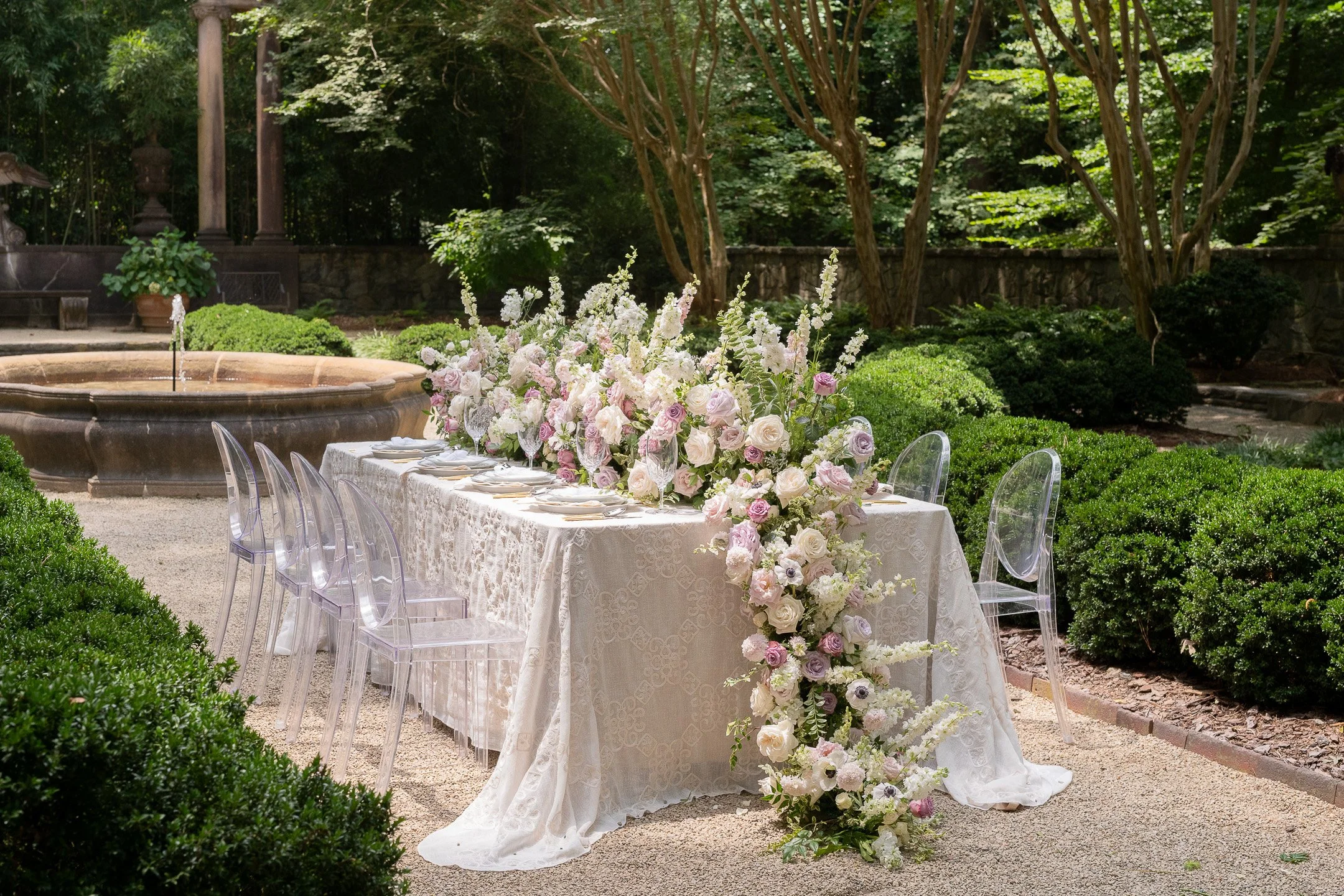 Elegant outdoor garden dining setup with a long table adorned with a white lace tablecloth and an abundance of pink and white flowers cascading down. Clear chairs are neatly arranged, and a stone fountain is visible in the background, surrounded by l