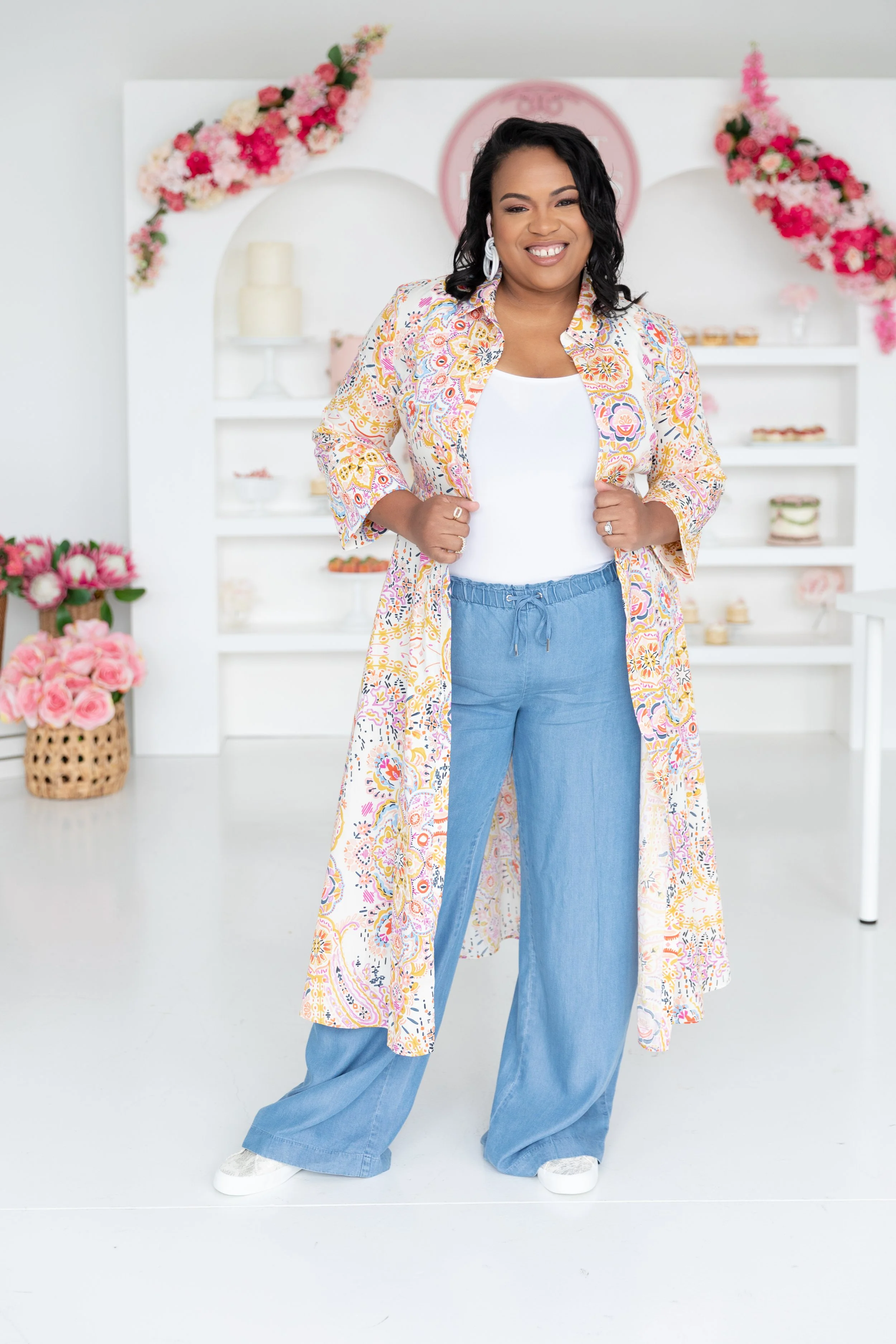 A woman standing in a bright room wearing a colorful long cardigan, white top, and blue pants, smiling at the camera. Background features floral decorations and shelves with decor items.