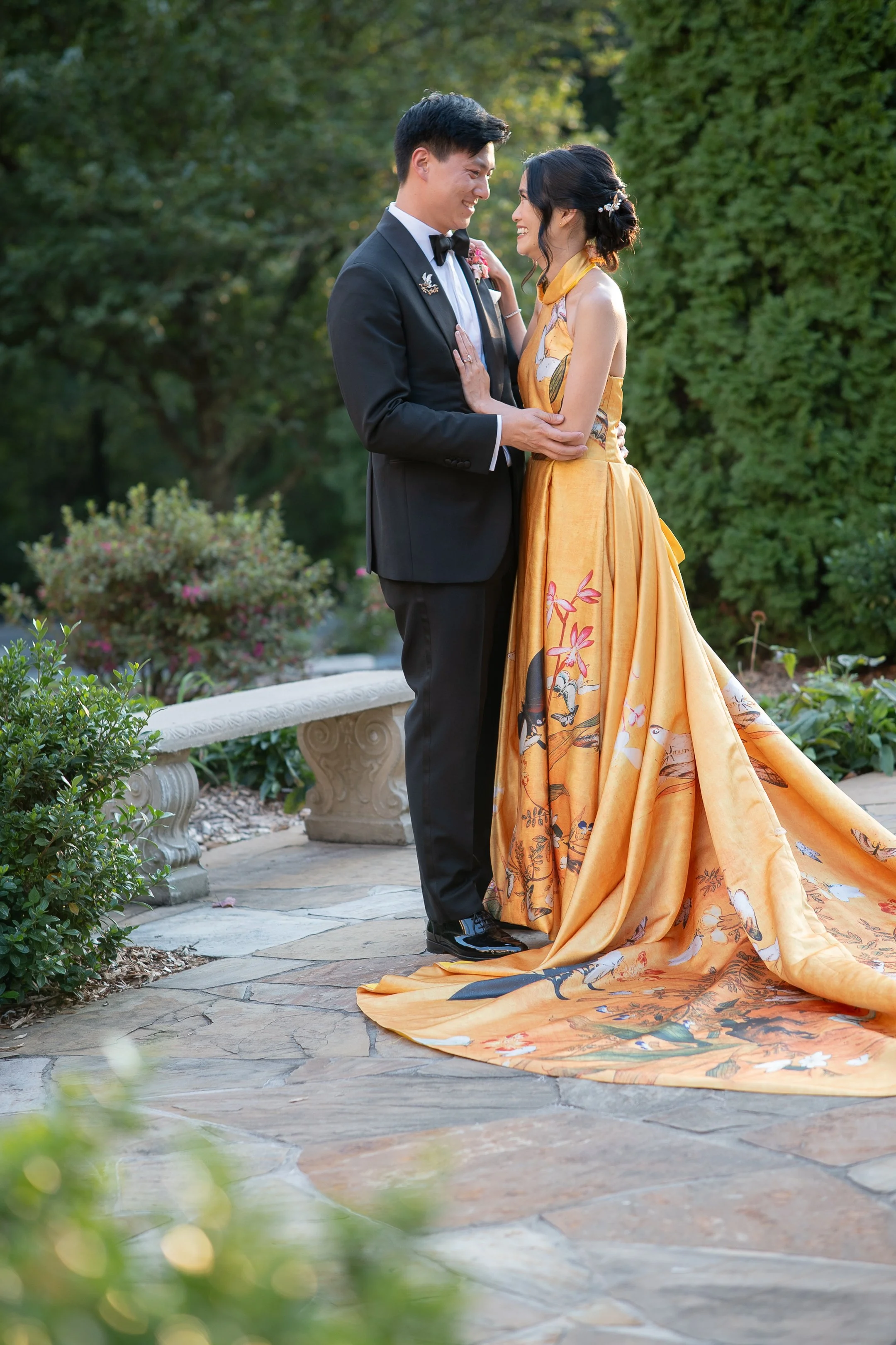 A couple dressed in formal attire stands close together in a garden, smiling at each other. The woman wears an elaborate, gold-colored gown with a long train decorated with floral and bird motifs. The man is in a black tuxedo with a bowtie.