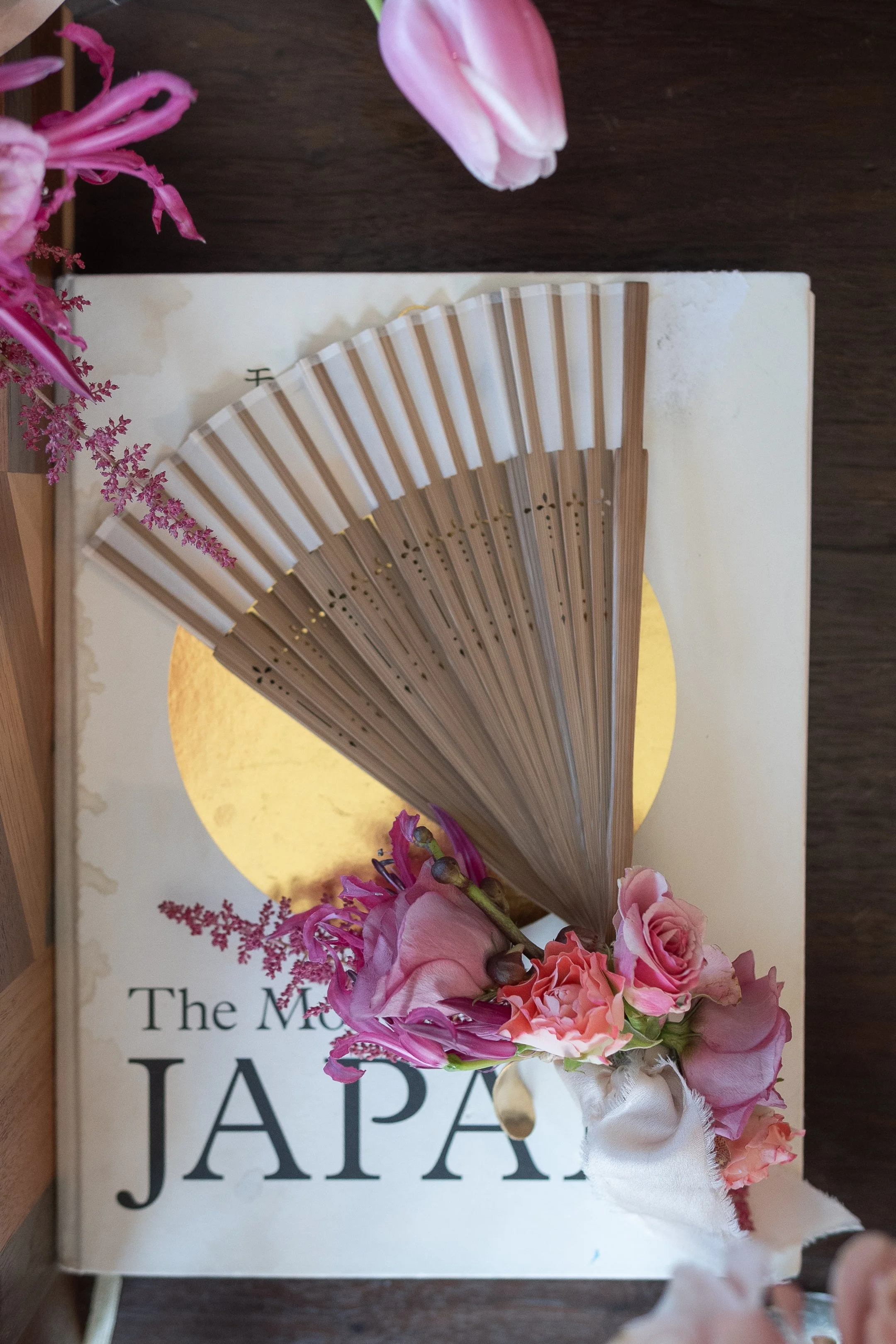 A Japanese fan rests on a book titled 'The Modern JAPA...' with a floral arrangement of pink roses, purple flowers, and a pink tulip, all placed on a dark wooden surface.