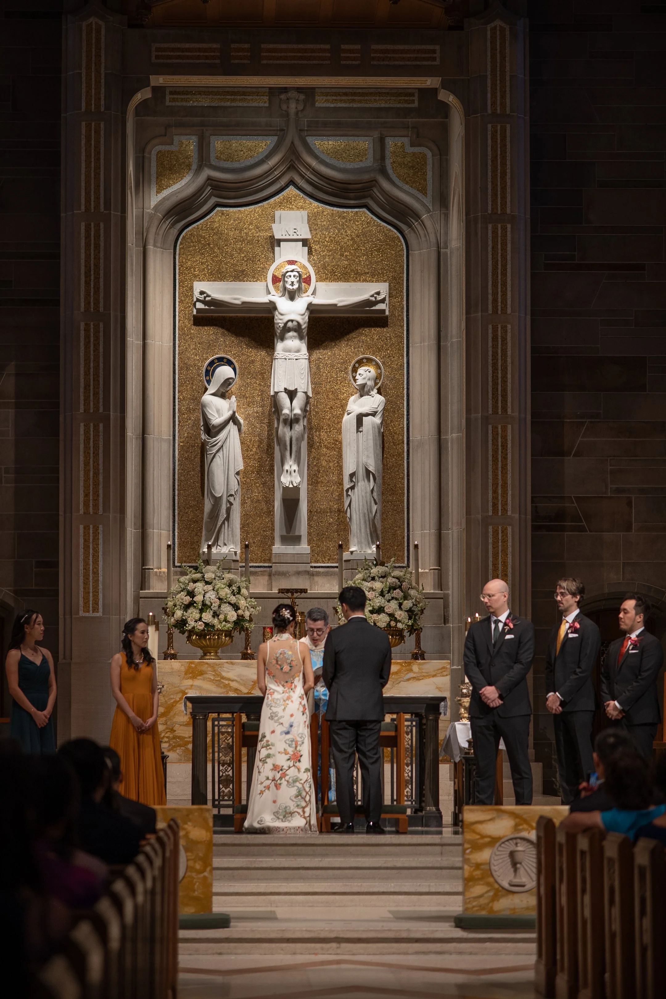 A wedding ceremony taking place in a church with a large crucifix and statues of Jesus, Mary, and John in the background. The bride and groom stand at the altar with a priest, and friends or family members are seated in the pews.