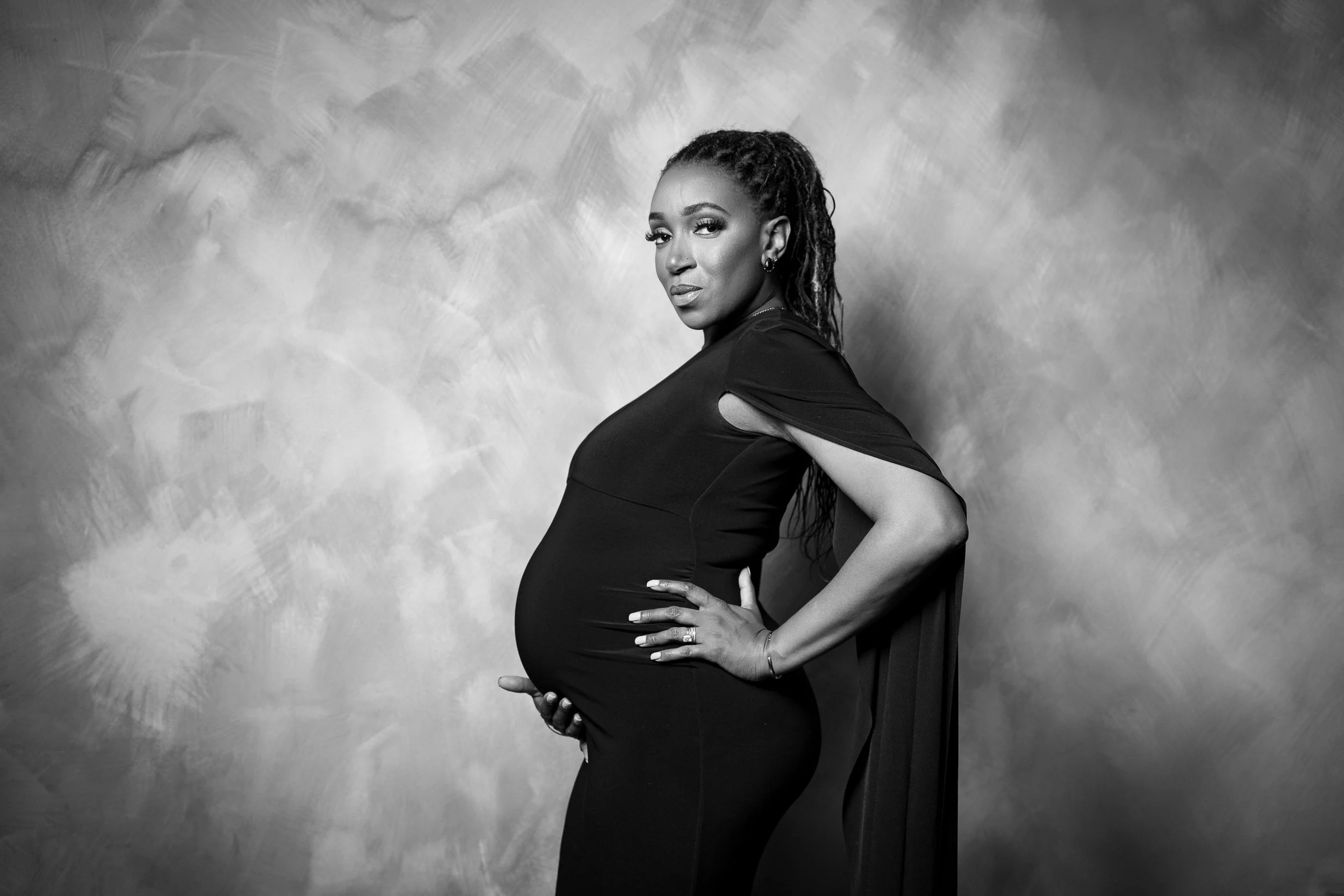 Black and white portrait of a pregnant woman standing against a textured background, wearing a fitted dress with cutouts on the sleeves, with her left hand on her hip and right hand on her belly, looking at the camera.