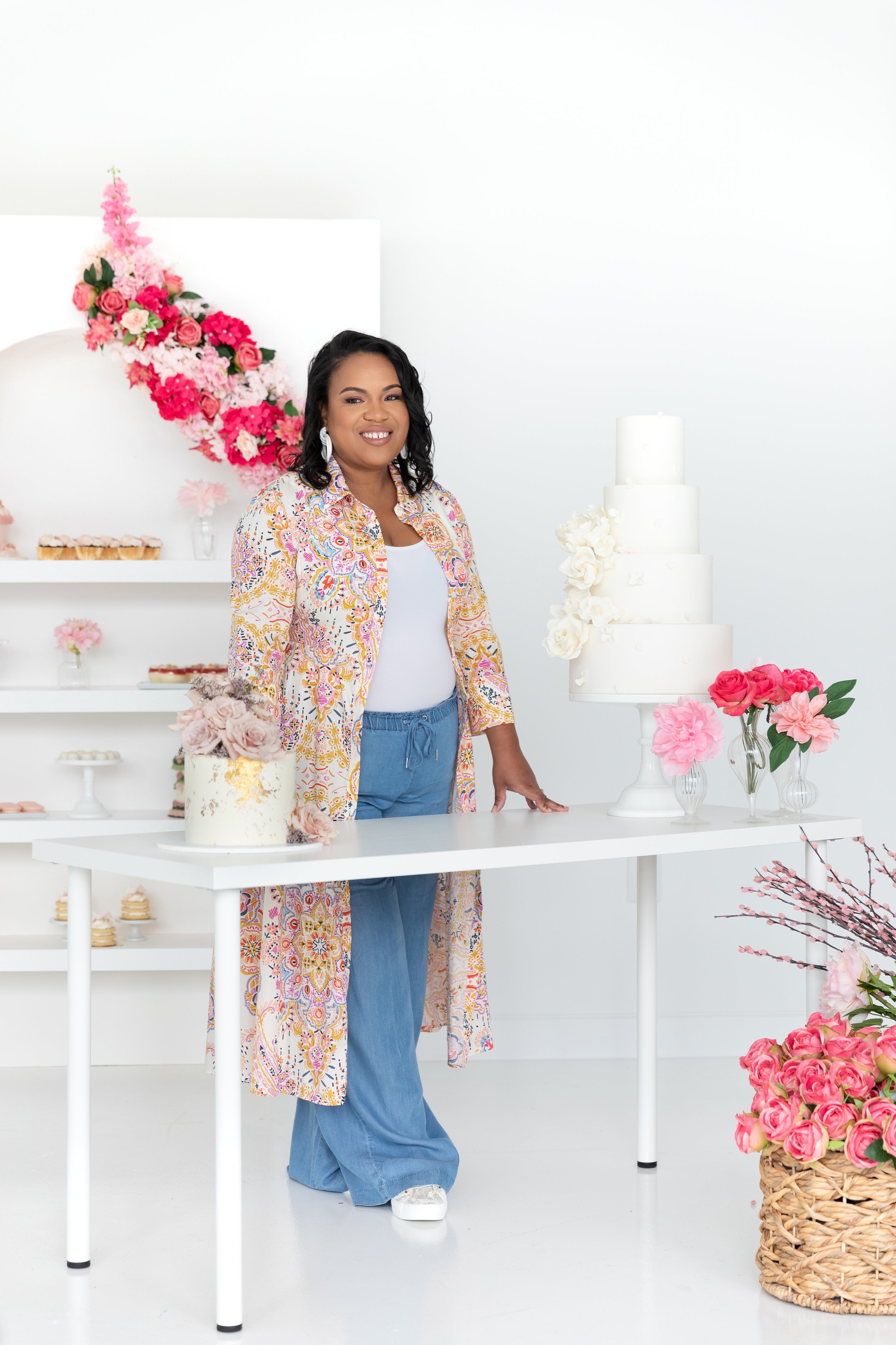 A woman standing by a table with a white tiered wedding cake and floral decorations. She's wearing a colorful long shirt, white top, and blue pants. The background features shelves with cupcakes and pastries.
