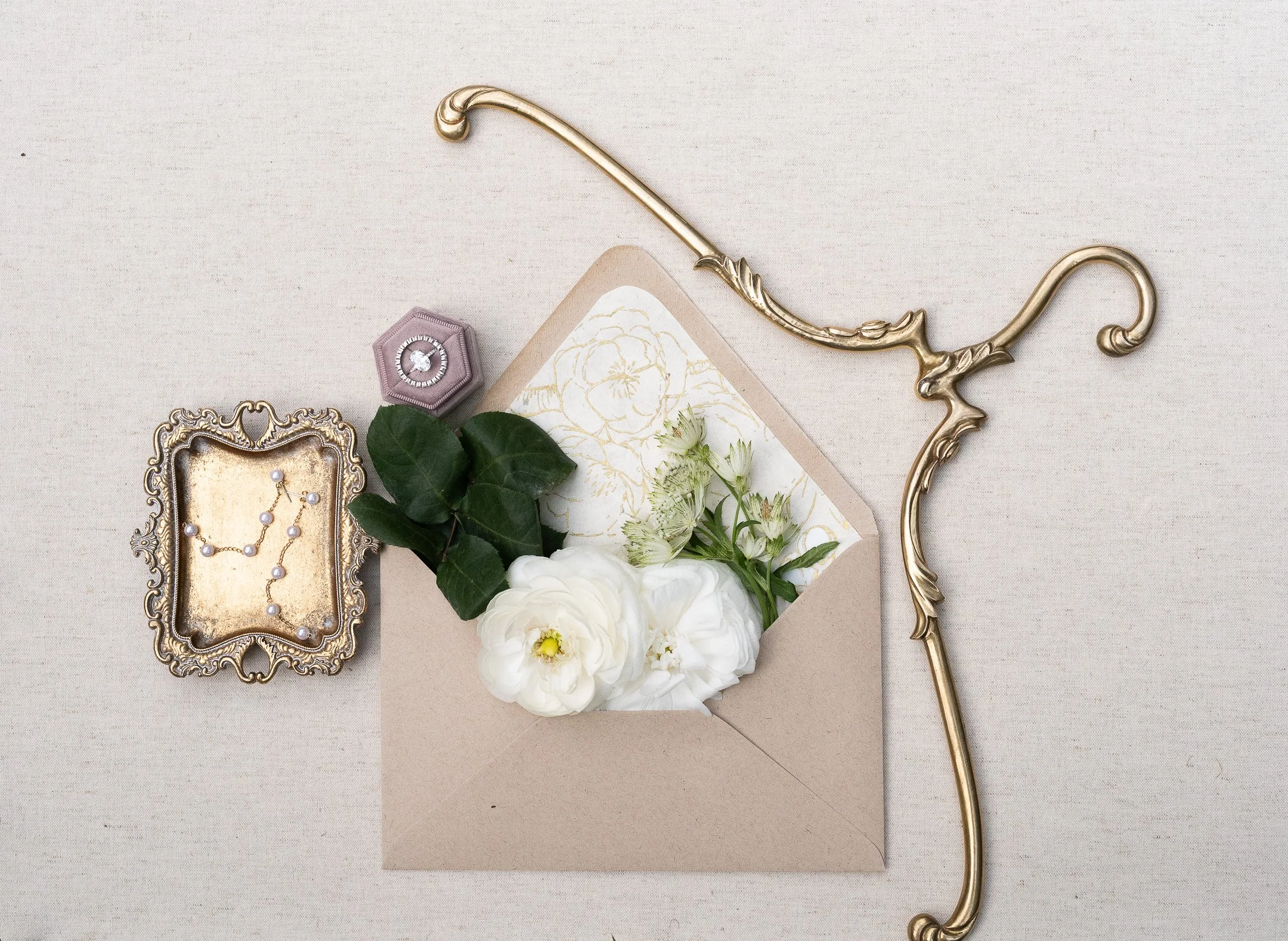 Flat lay of a decorative invitation with flowers, a vintage tray holding a pearl necklace, a hexagonal ring box, and ornate gold embellishments on a beige background.