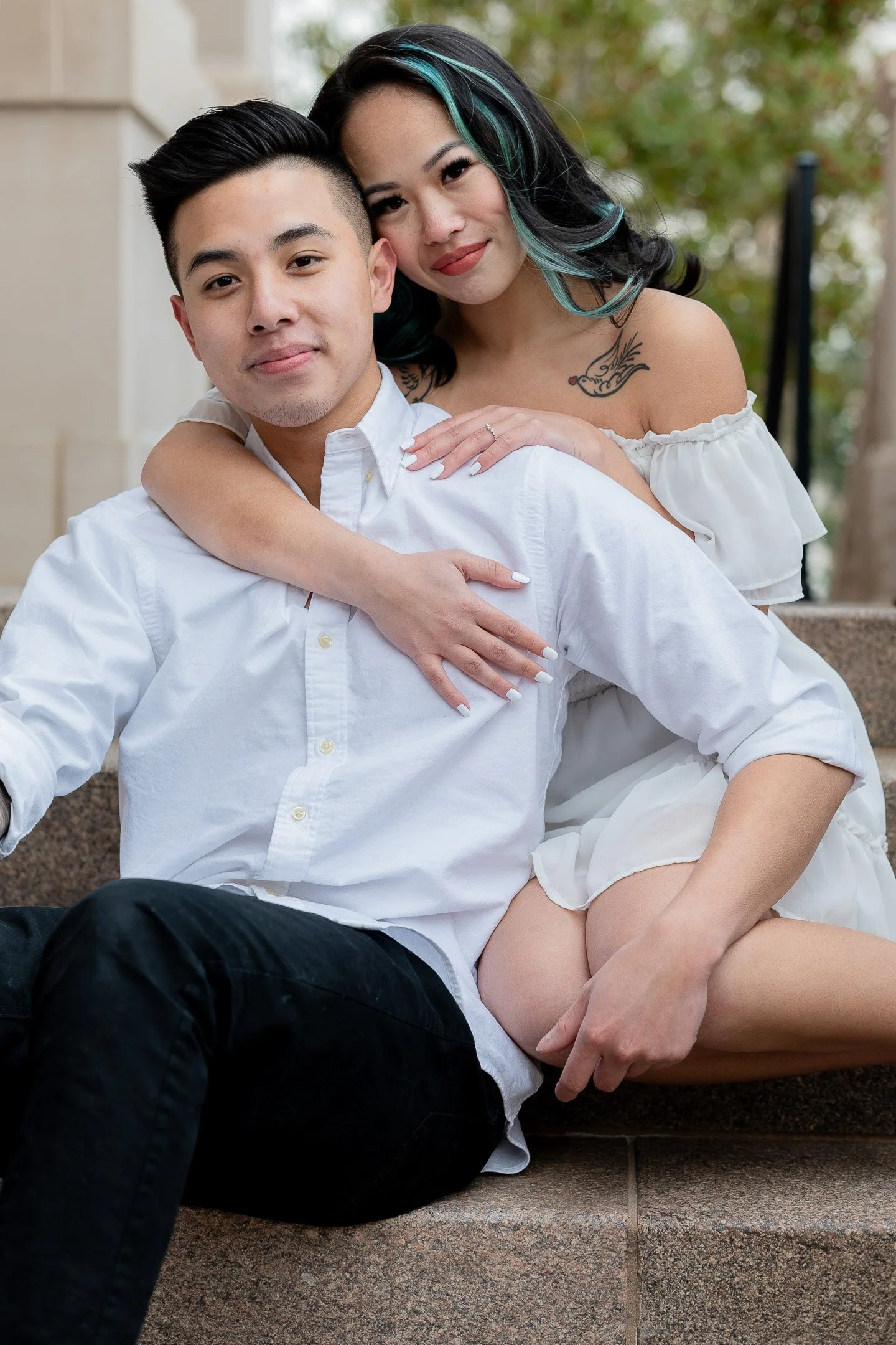 A couple sitting on outdoor steps, with the woman wrapping her arms around the man's shoulders, both smiling.