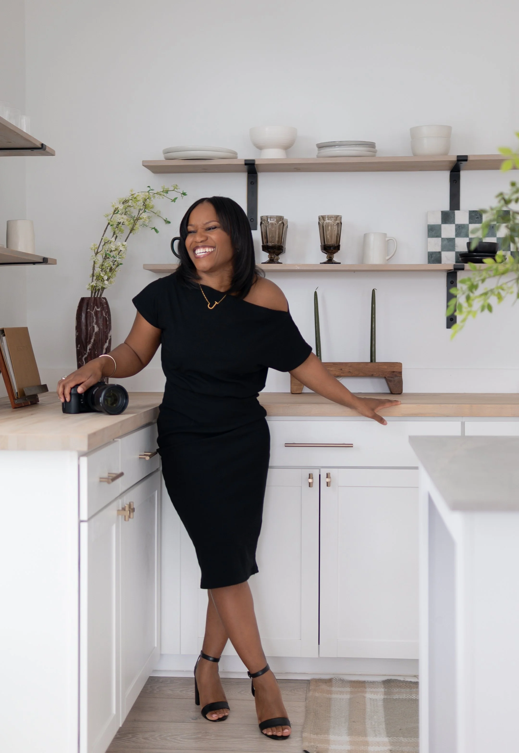 Woman in a kitchen wearing a black dress, smiling and leaning against a counter with a camera on it.