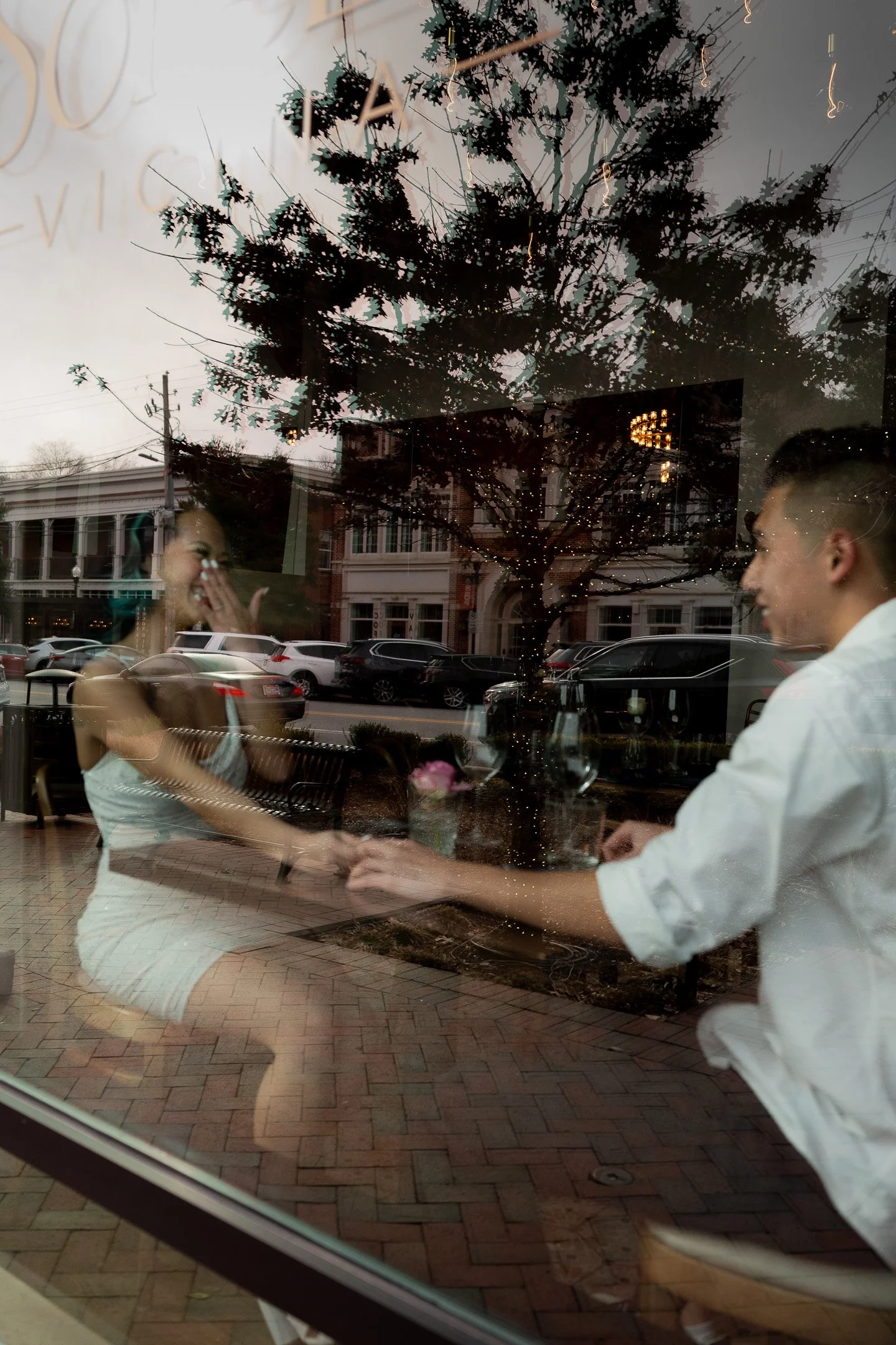 A couple sits at a table in a cafe, holding hands and smiling, with the outside street scene reflected in the window.
