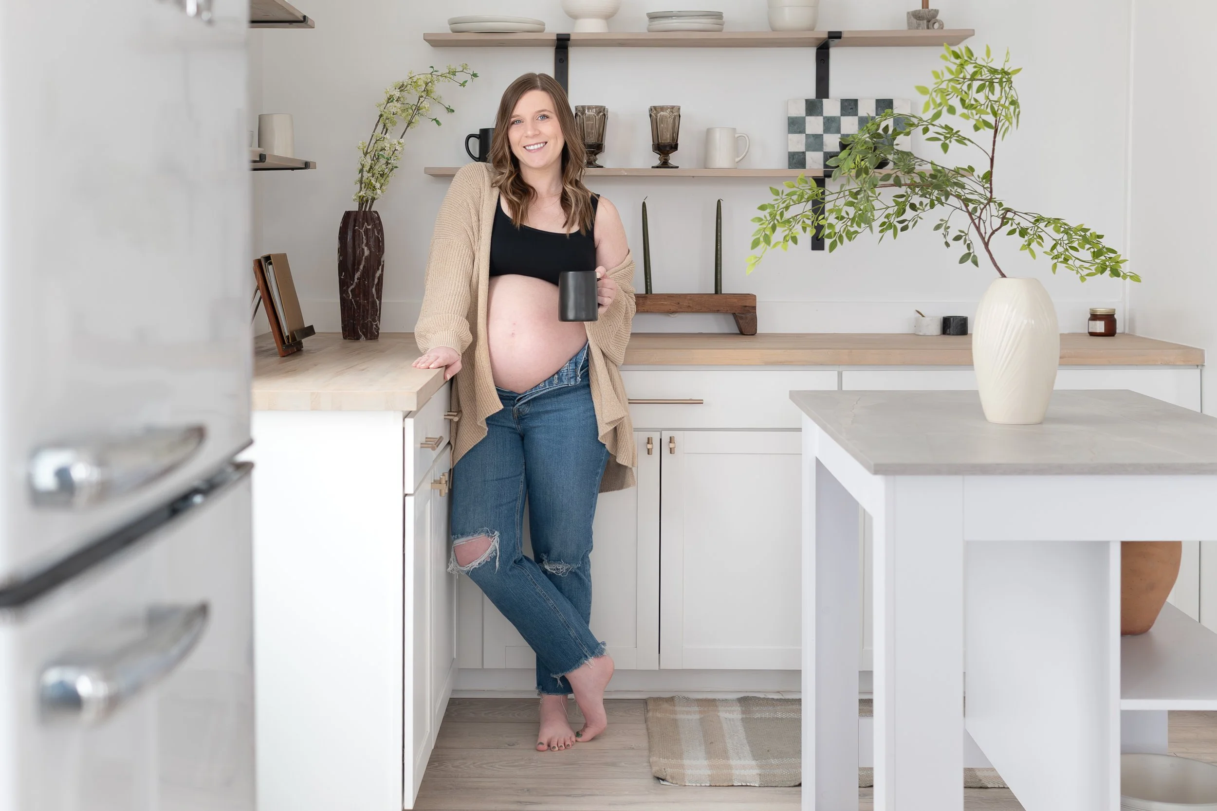 Pregnant woman in a kitchen holding a mug, wearing a black top, beige cardigan, and ripped jeans; standing by a countertop with a vase and potted plant.
