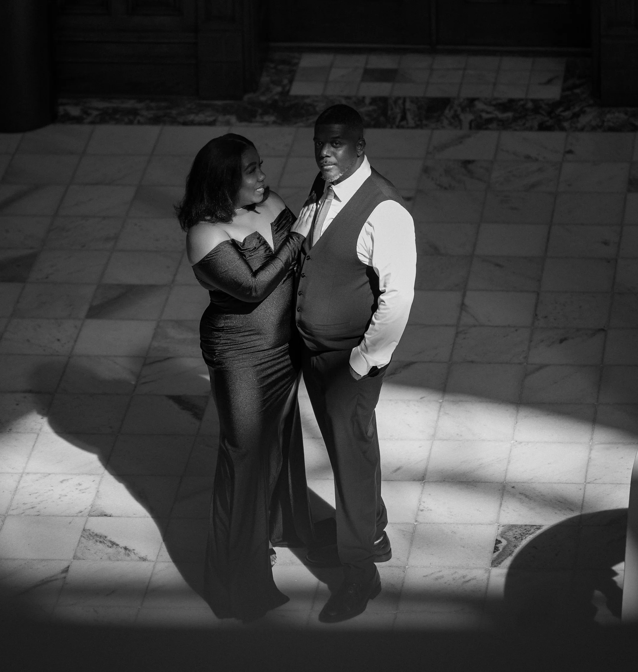 A black and white photo of a woman and a man standing close together on a tiled floor, with a spotlight focused on them. The woman is wearing a long, off-shoulder dress and gloves, and is looking at the man. The man is wearing a vest and tie, looking