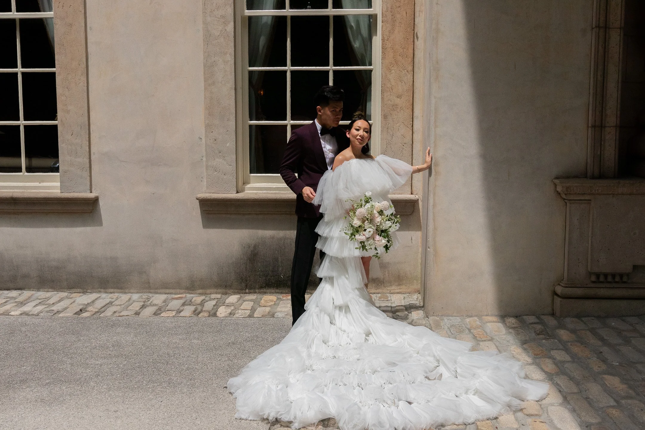 Bride and groom standing outside by a wall and window; bride wearing a white dress with a long train holding a bouquet.