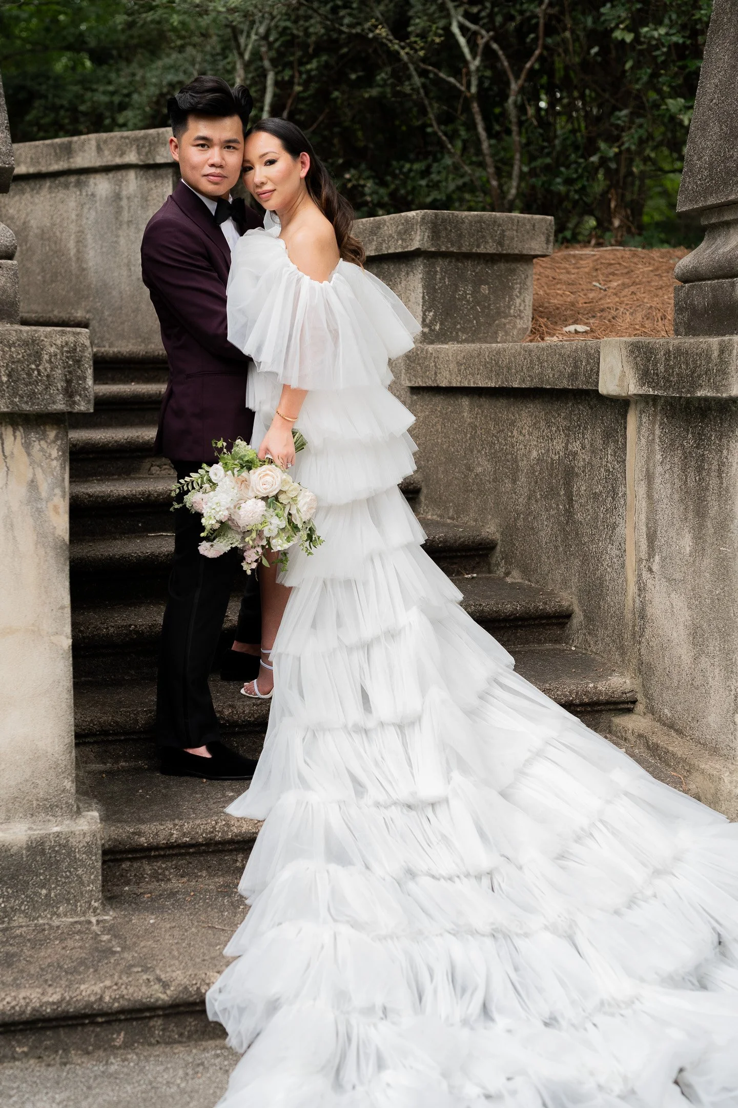 A bride and groom posing on stone steps. The bride wears a white, tiered, off-the-shoulder gown and holds a bouquet. The groom is in a dark suit, embracing the bride.