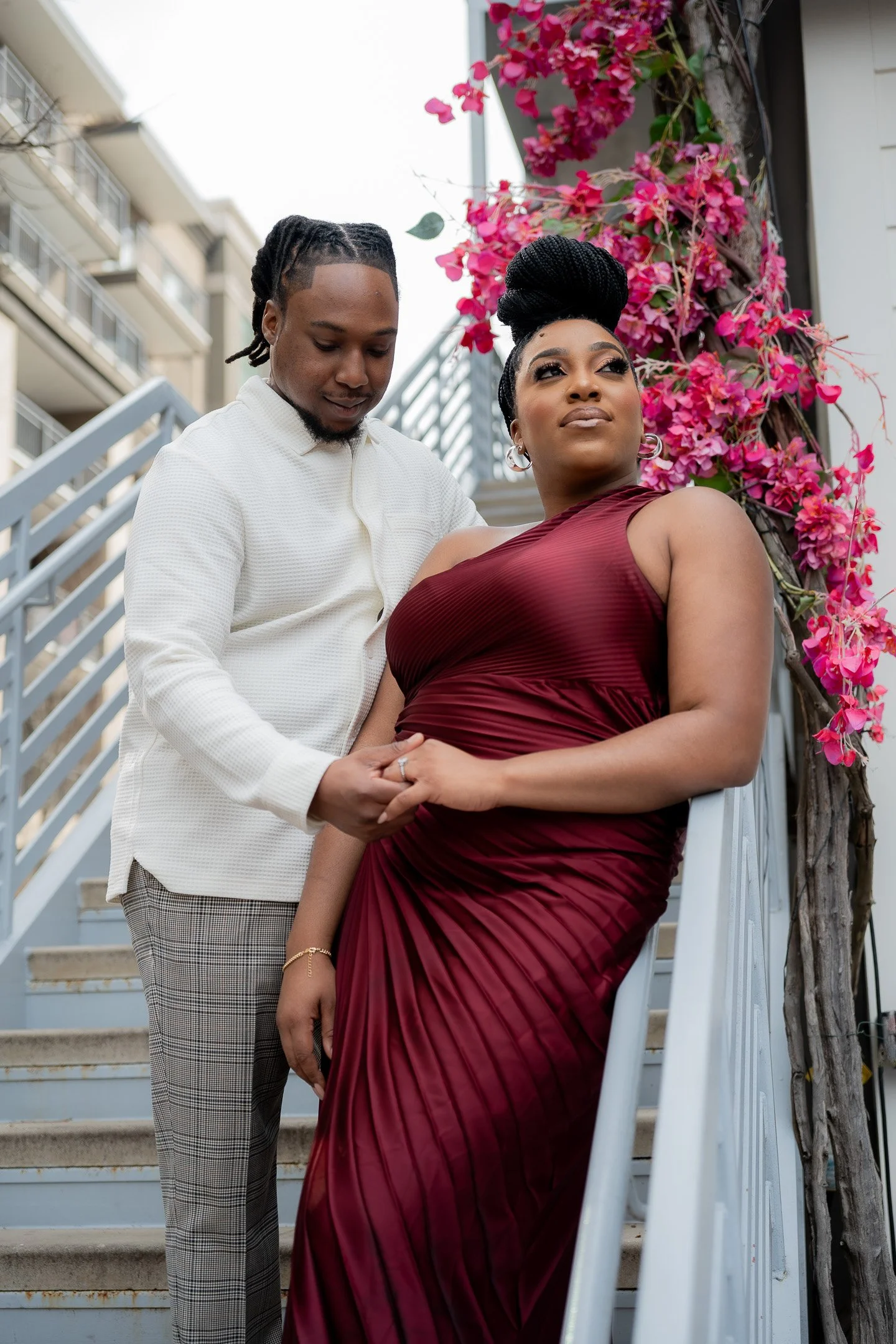 A couple poses on an outdoor staircase adorned with pink flowers. The woman is wearing a burgundy gown, and the man is in a white shirt and plaid pants, holding her hand tenderly.