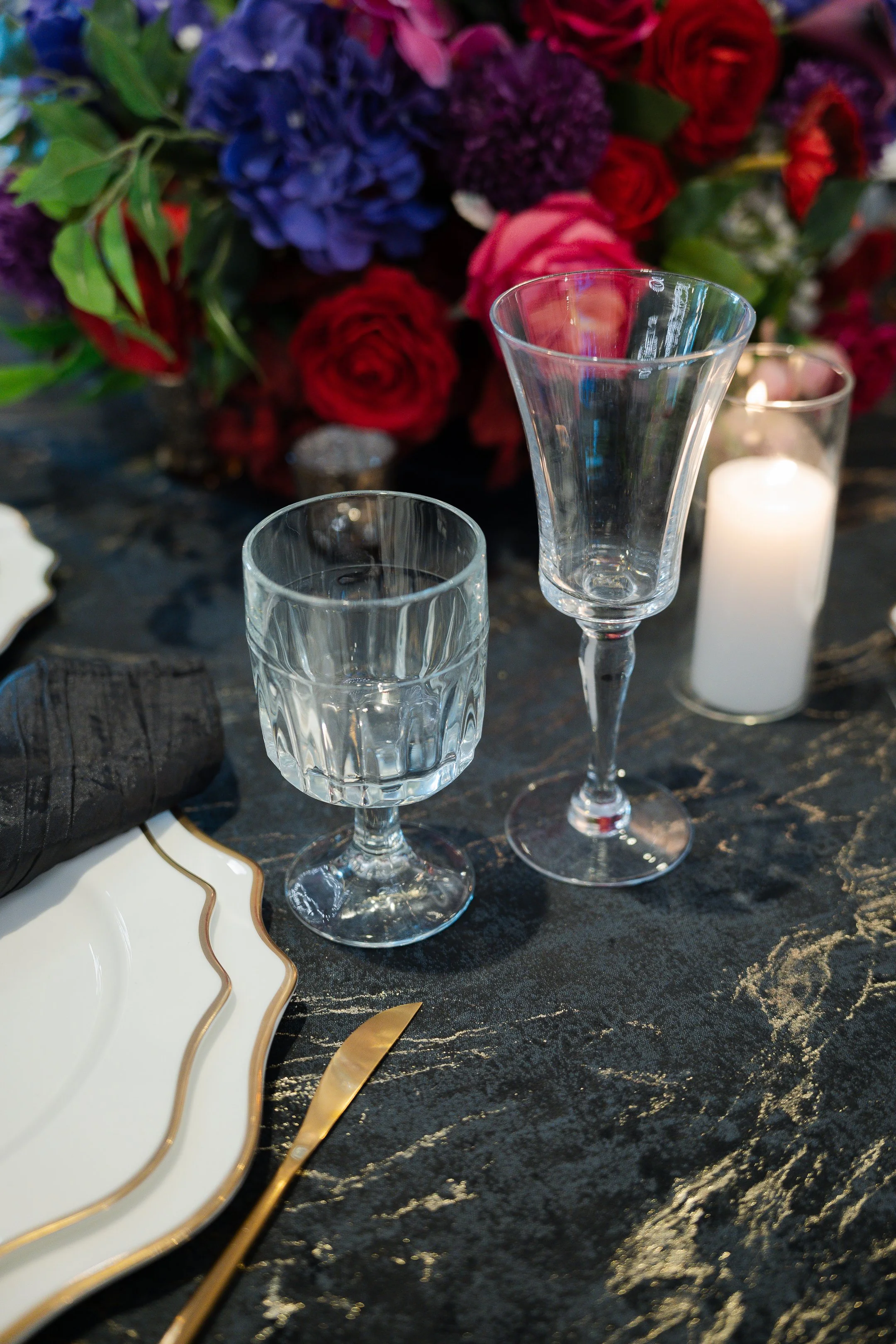 Elegant table setting with two crystal glasses, a white plate with gold trim, black napkin, gold-toned knife, lit candle, and a large bouquet of red, purple, pink, and green flowers on a dark marbled table.