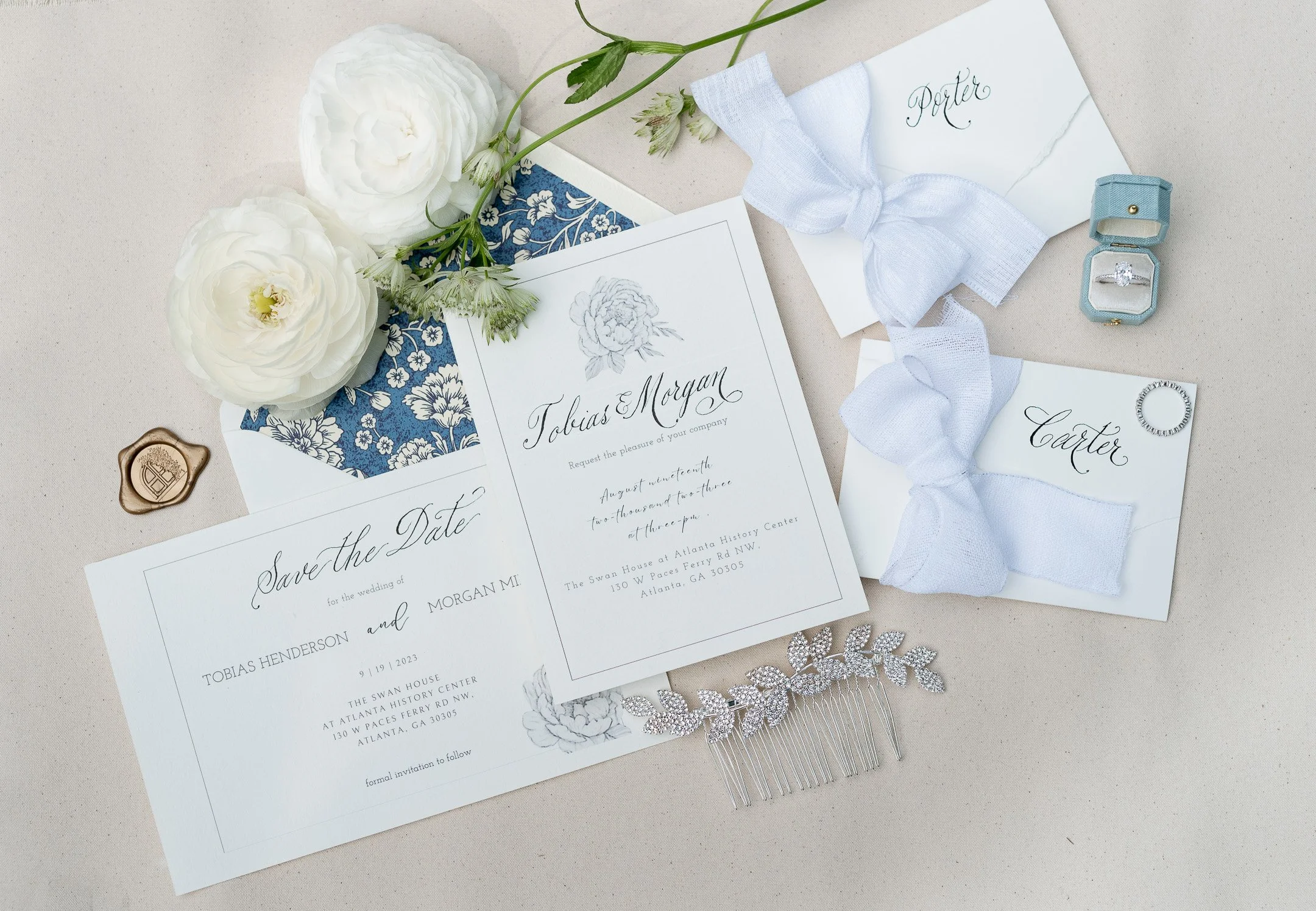 Wedding invitation set with elegant script, white flowers, decorative envelope, seal, ring box, ribbon, and hair accessory on a beige surface.