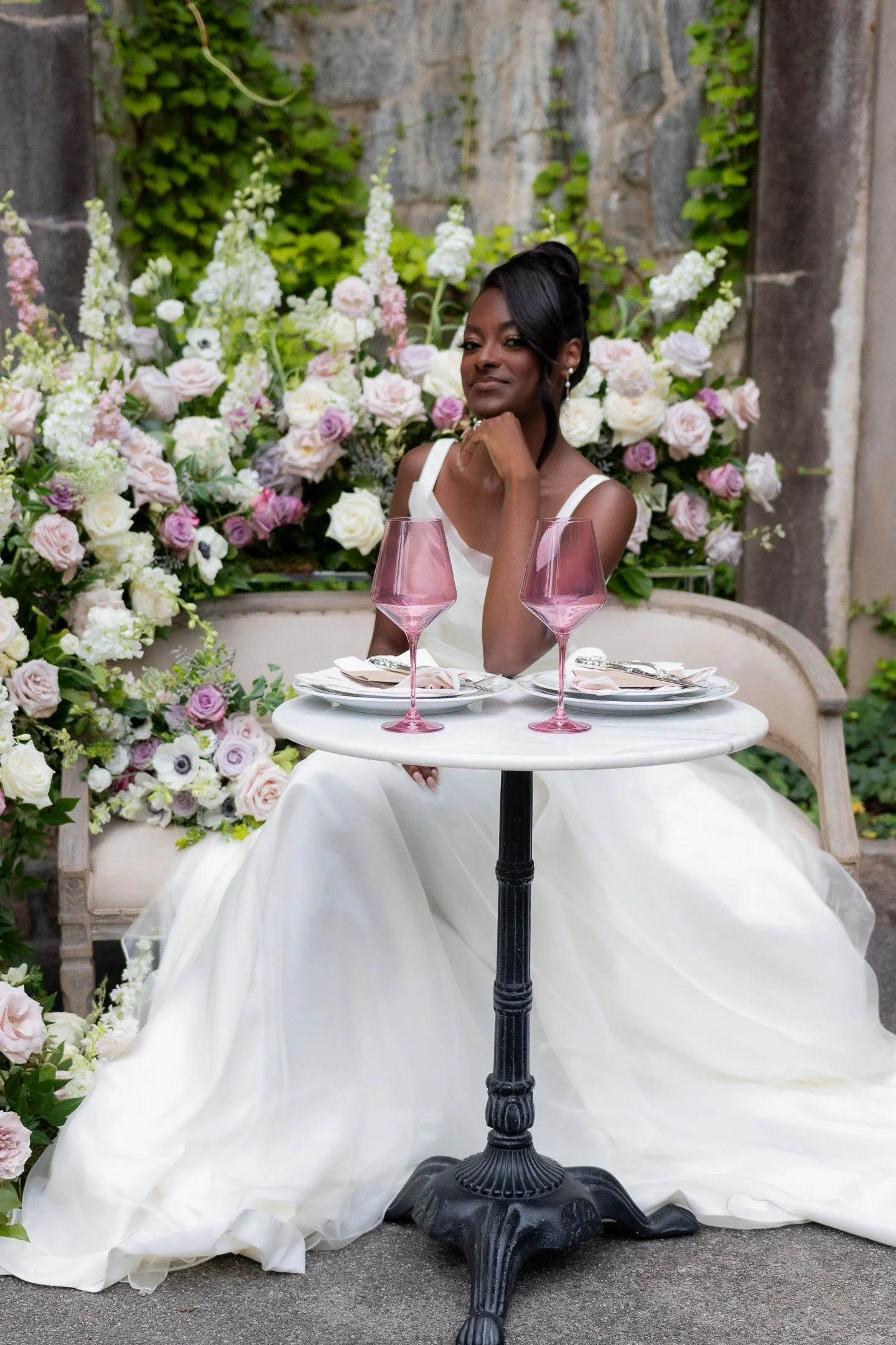 Bride in wedding gown sitting at a small round table outdoors, surrounded by floral arrangements, with two pink wine glasses on the table.