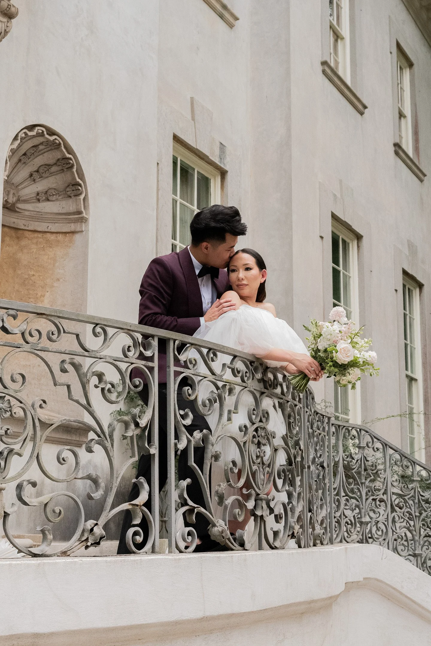 A couple stands on a decorative iron balcony. The man in a suit kisses the woman's forehead. She is holding a bouquet of flowers.