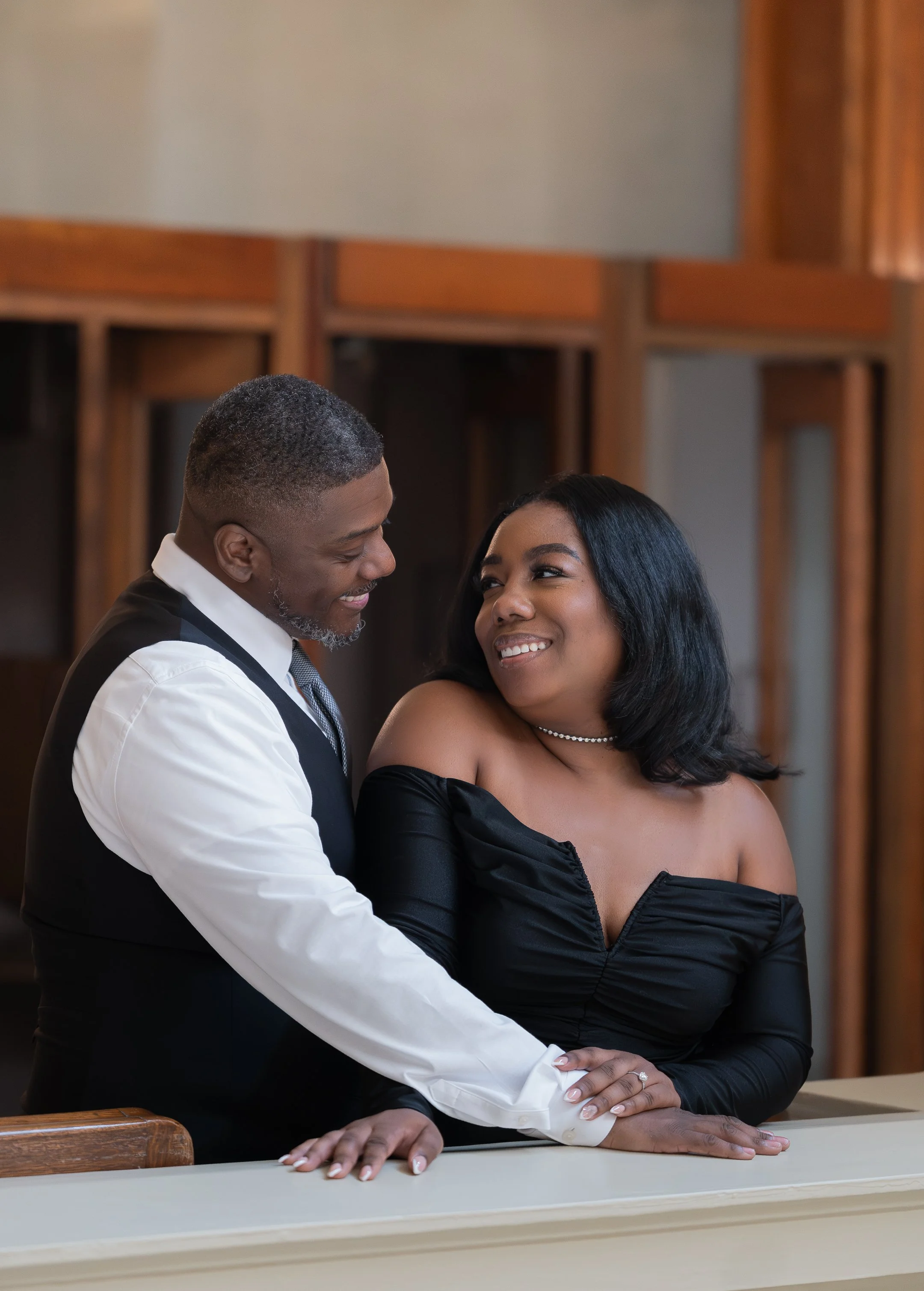 A couple at their wedding ceremony, smiling and looking into each other's eyes, with a wooden background.