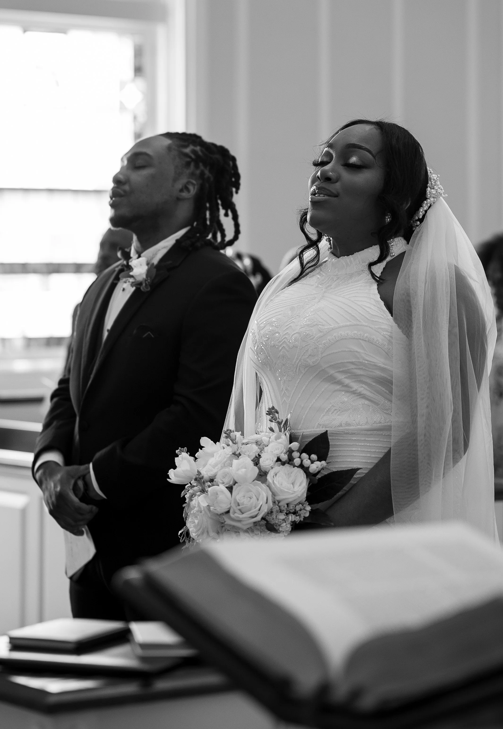 A black and white photo of a bride and groom standing in a church. The bride is wearing a wedding gown with a veil and holding a bouquet of flowers. The groom is in a suit with a bow tie. A book, possibly a Bible, appears in the foreground.