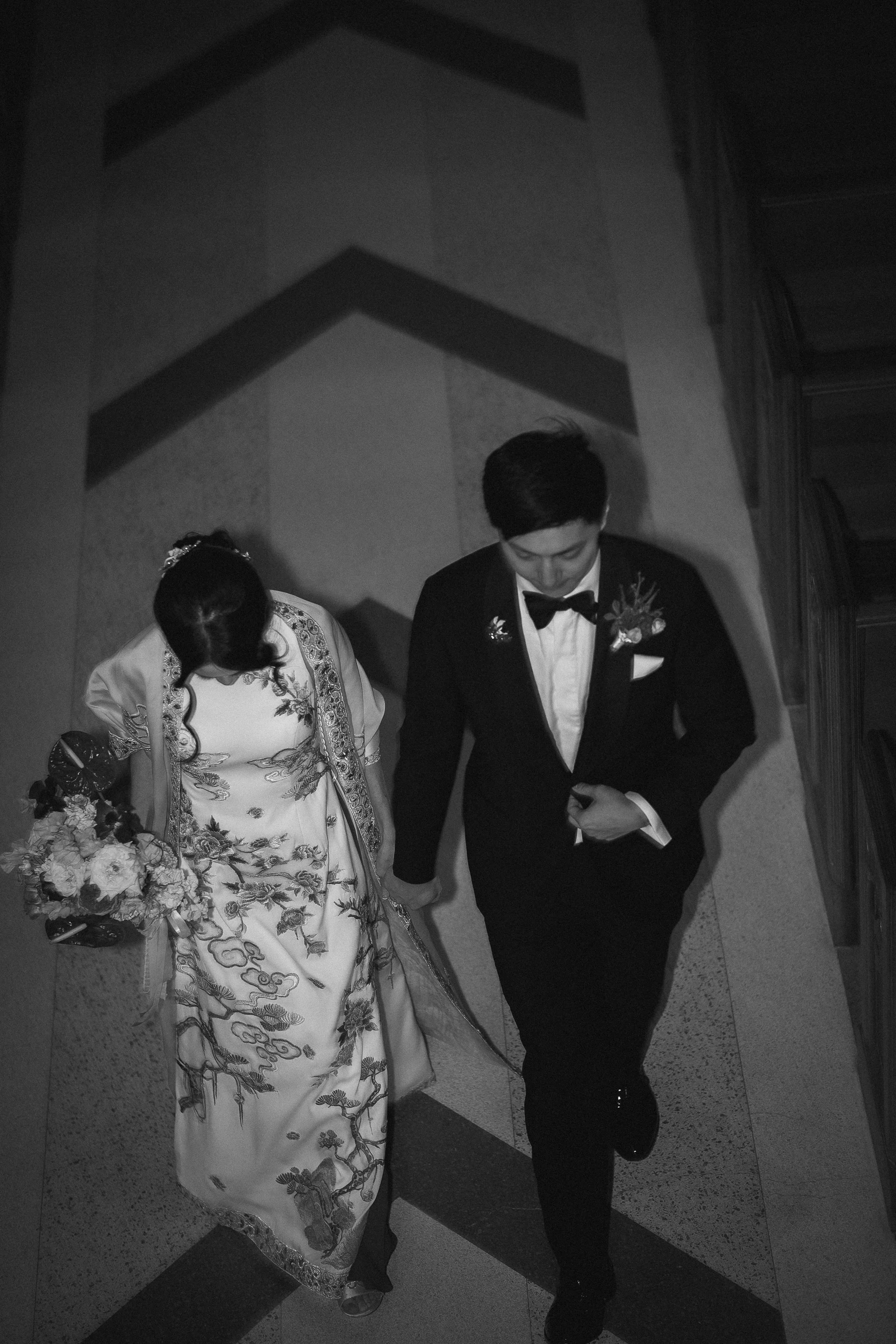 Black and white photo of a bride and groom walking hand in hand down a staircase, viewed from above. The bride is wearing a traditional wedding dress with floral embroidery and carrying a bouquet, while the groom is dressed in a tuxedo with a bow tie